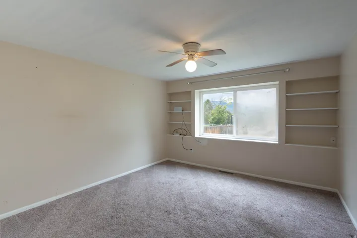 Neutral-toned room featuring a large window, built-in shelving, a ceiling fan,  and carpet flooring