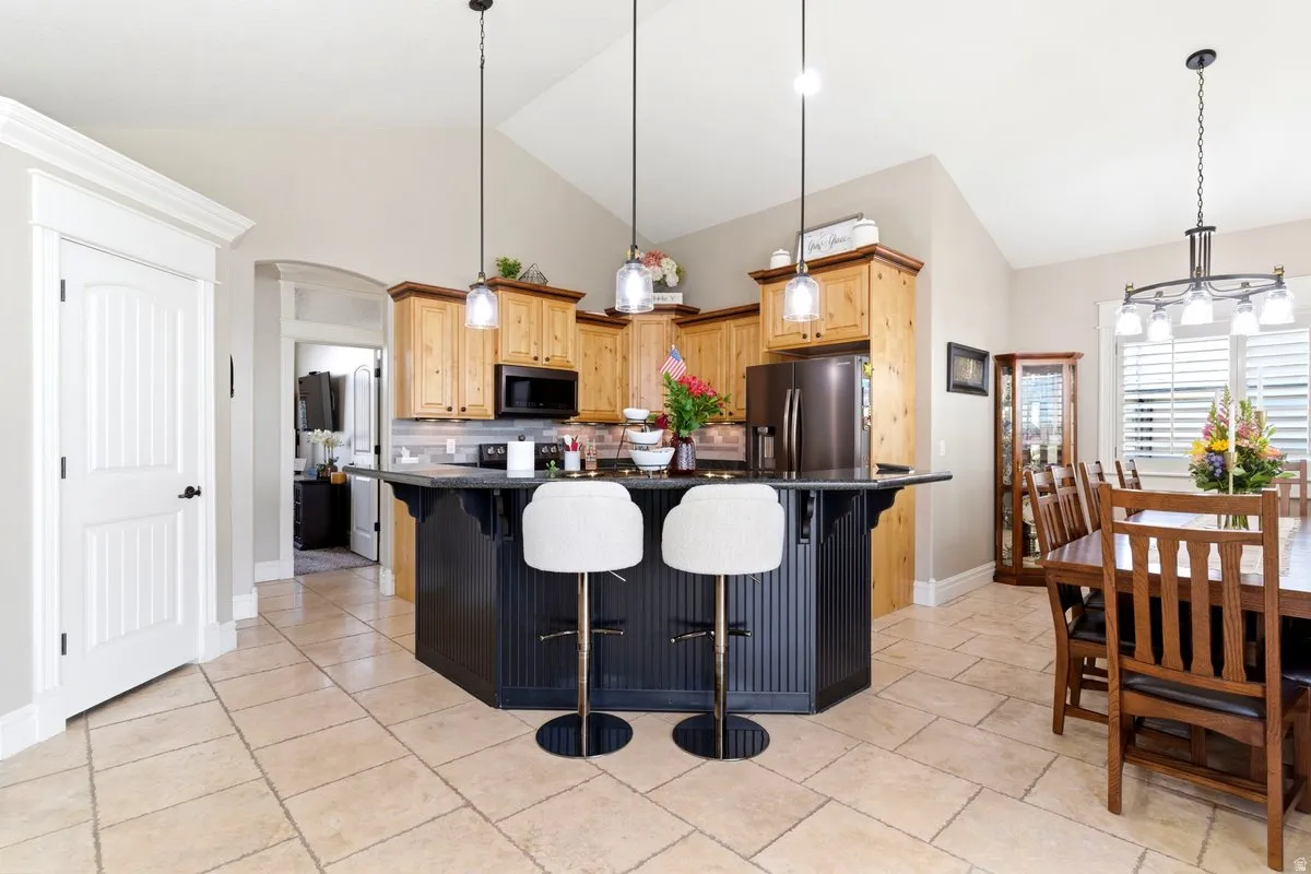 Open-concept kitchen and dining area featuring a dark-toned island with beadboard paneling, light wood cabinetry, stainless steel appliances, a tile backsplash, and light-toned tile flooring