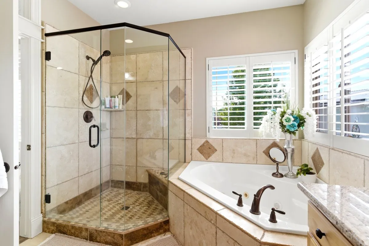Bathroom featuring a corner soaking tub with bronze fixtures, a glass-enclosed shower with tiled walls and floor, and plantation shutters on the window