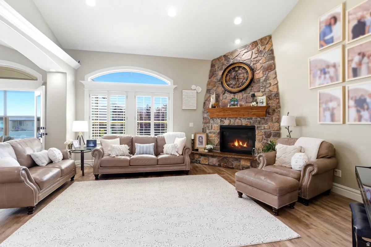Vaulted ceiling living space featuring a stone-clad fireplace with wood mantel, large arched windows with plantation shutters, wood-finish flooring, and recessed lighting