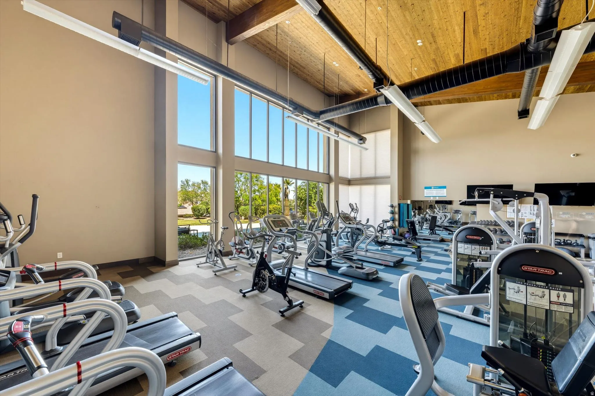 Spacious fitness center featuring high ceilings with exposed wood beams, ample natural light from extensive window walls, patterned carpet flooring, and a variety of exercise equipment