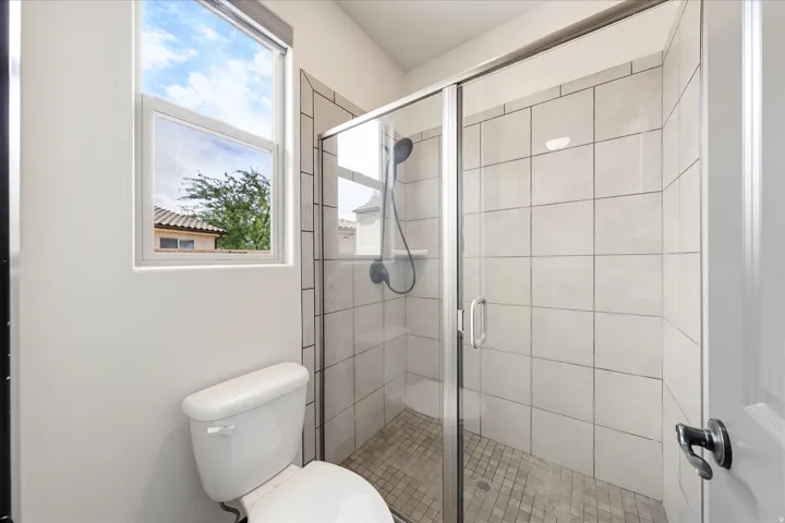 Glass-enclosed shower with subway tile surround and mosaic tile flooring, complemented by a white toilet and a window with a roller shade