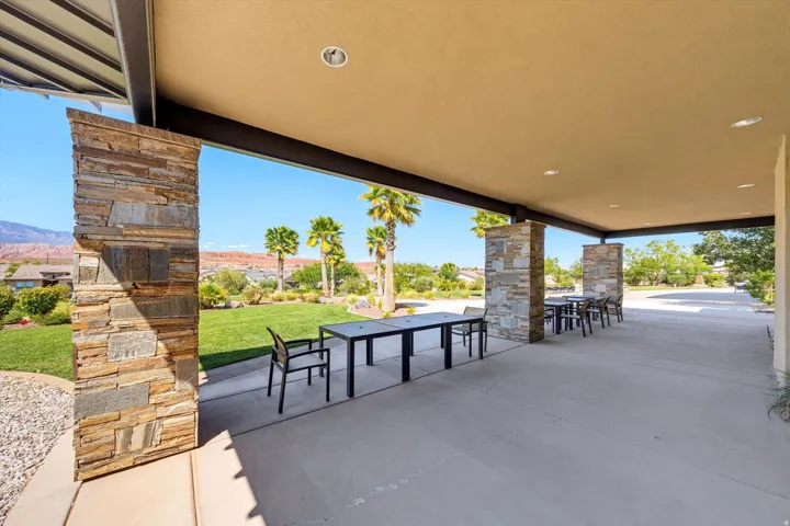Expansive covered patio featuring stone-clad pillars, recessed lighting, concrete flooring, and a verdant lawn beyond