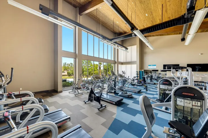 Spacious fitness center featuring high ceilings with exposed wood beams, ample natural light from extensive window walls, patterned carpet flooring, and a variety of exercise equipment