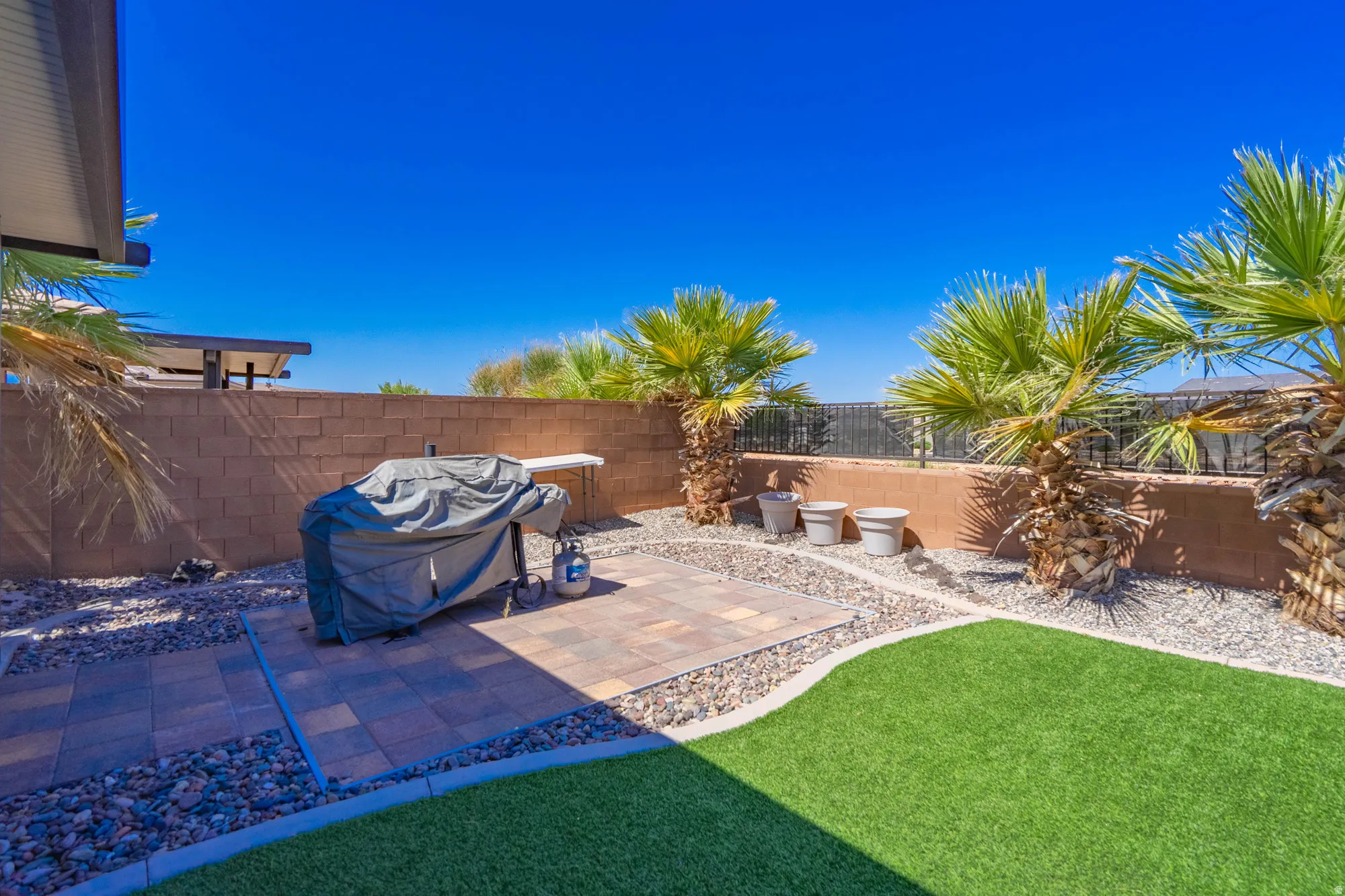 Backyard featuring artificial turf, a paver patio, a perimeter block wall, decorative rock landscaping, and mature palm trees