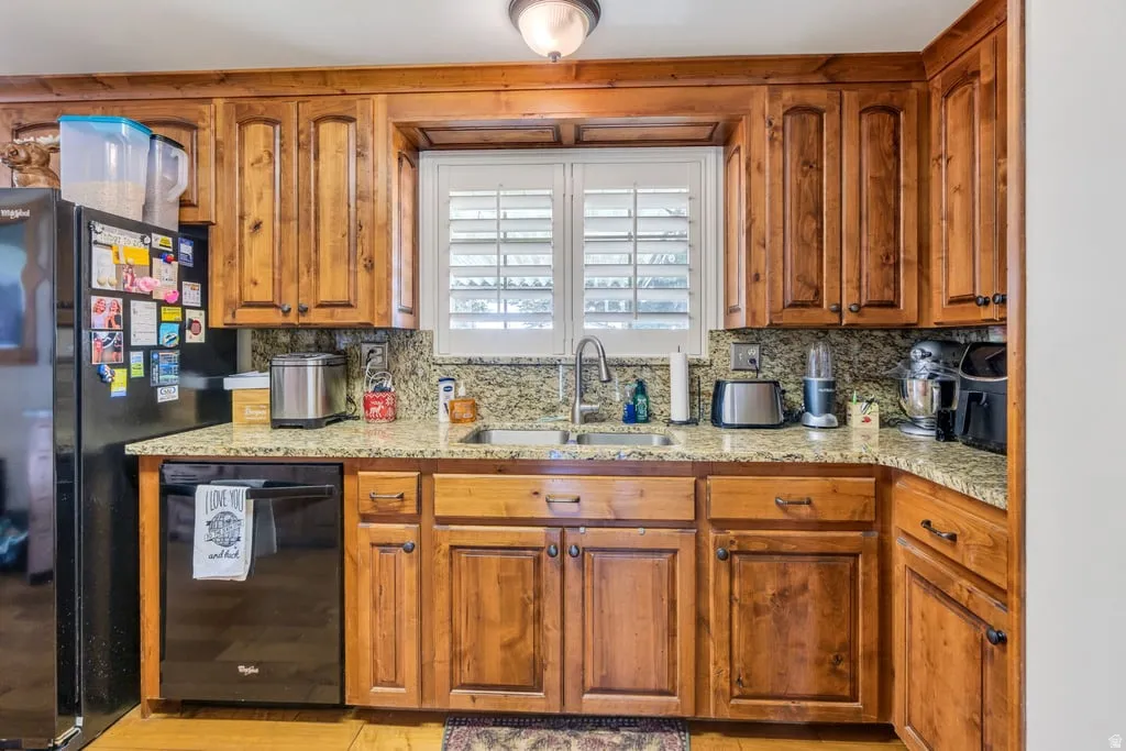 Kitchen featuring wood cabinetry, light-toned granite countertops, a double basin sink, black appliances, and a window with white louvered shutters