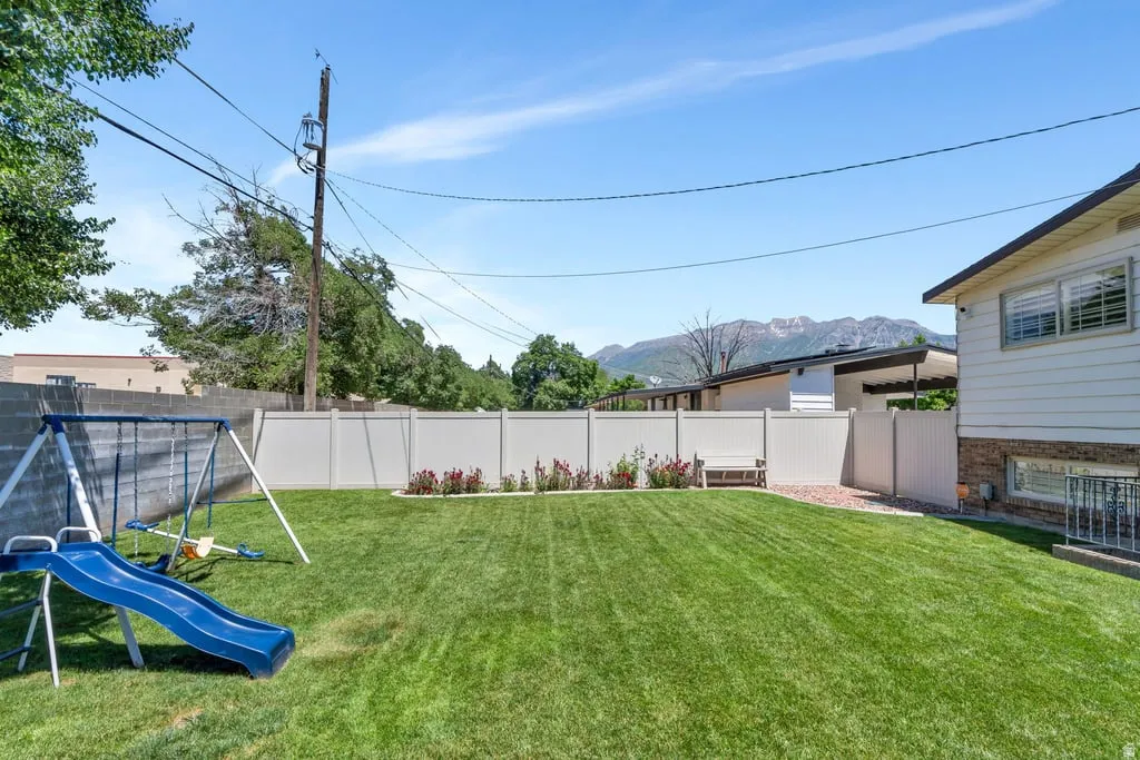 Expansive backyard featuring a manicured lawn, white vinyl fencing, a defined flower bed with plantings, and a partial mountain backdrop