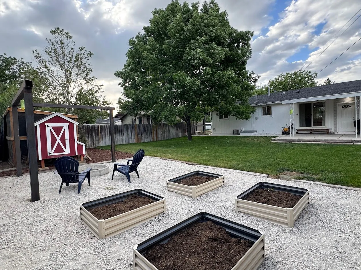 Expansive backyard with a large shade tree, multiple raised garden beds, and a pea gravel patio featuring a built-in fire pit