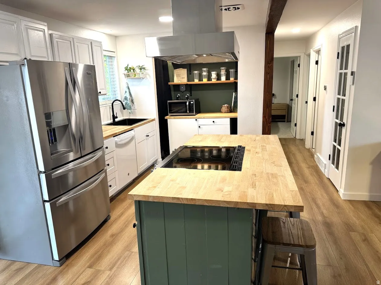 Modern kitchen featuring a central island with an integrated cooktop, butcher block countertops, white cabinetry, stainless steel appliances, and wood-finish flooring