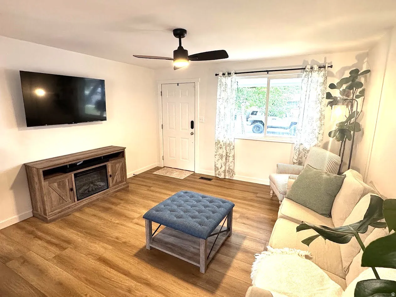 Living area featuring wood-finish flooring, a white entry door, and a ceiling fan with integrated lighting