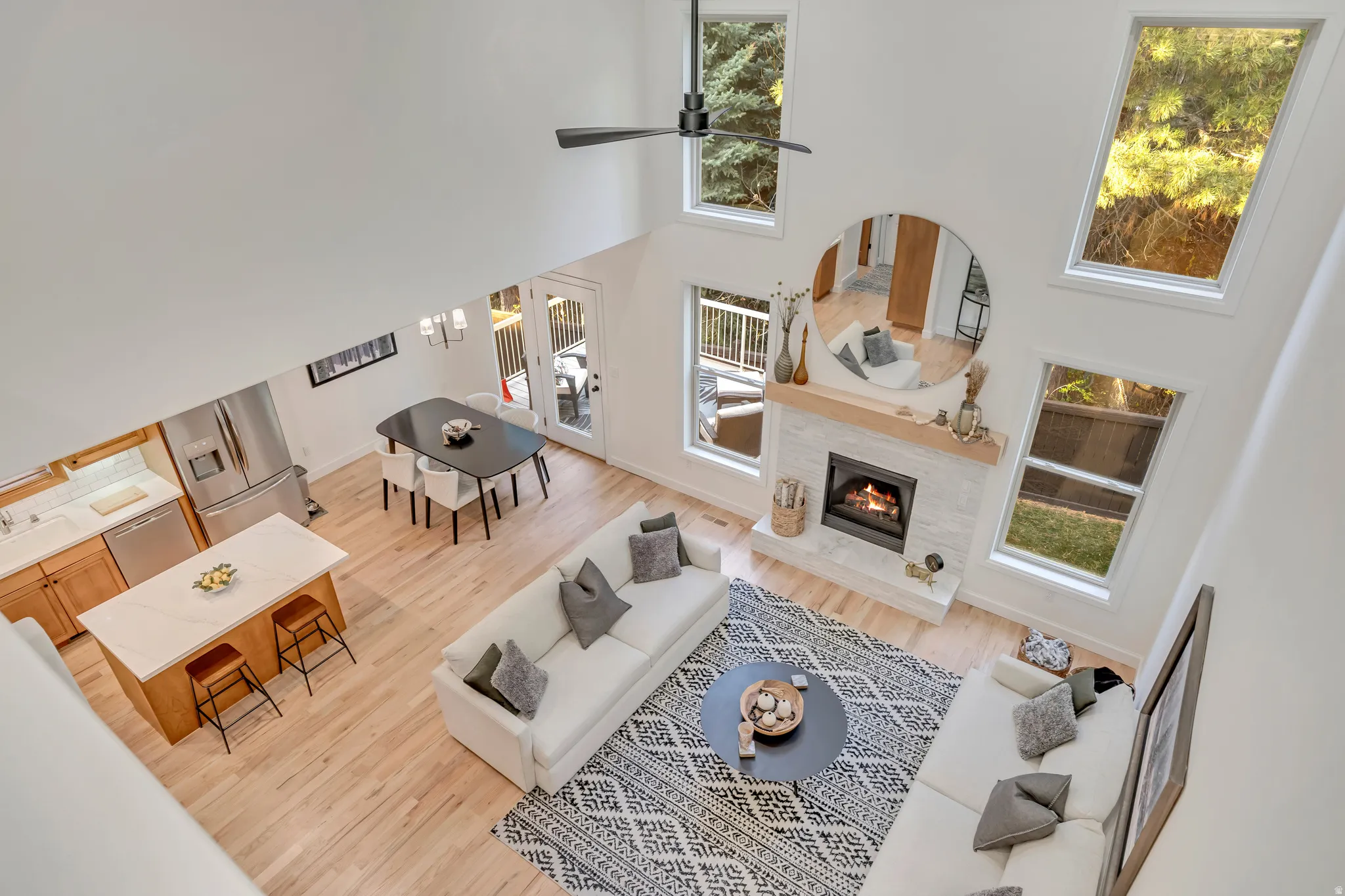 Open-concept living space featuring vaulted ceilings, abundant natural light from multiple windows, a modern fireplace with a light-toned tile surround and wood mantle, light wood-finish flooring, and a contemporary kitchen with a central island and stain
