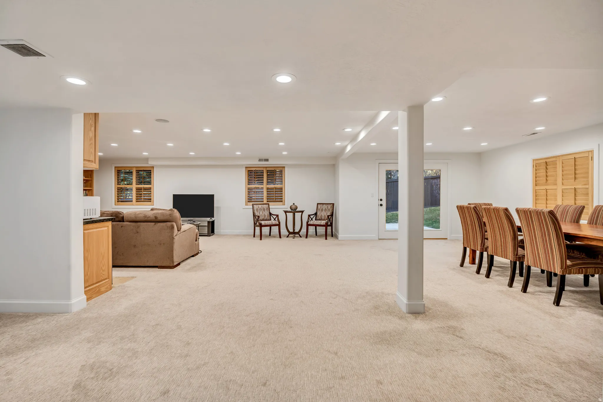 Expansive carpeted lower level featuring recessed lighting, multiple windows with wooden plantation shutters, and a glass-paneled door opening to the exterior