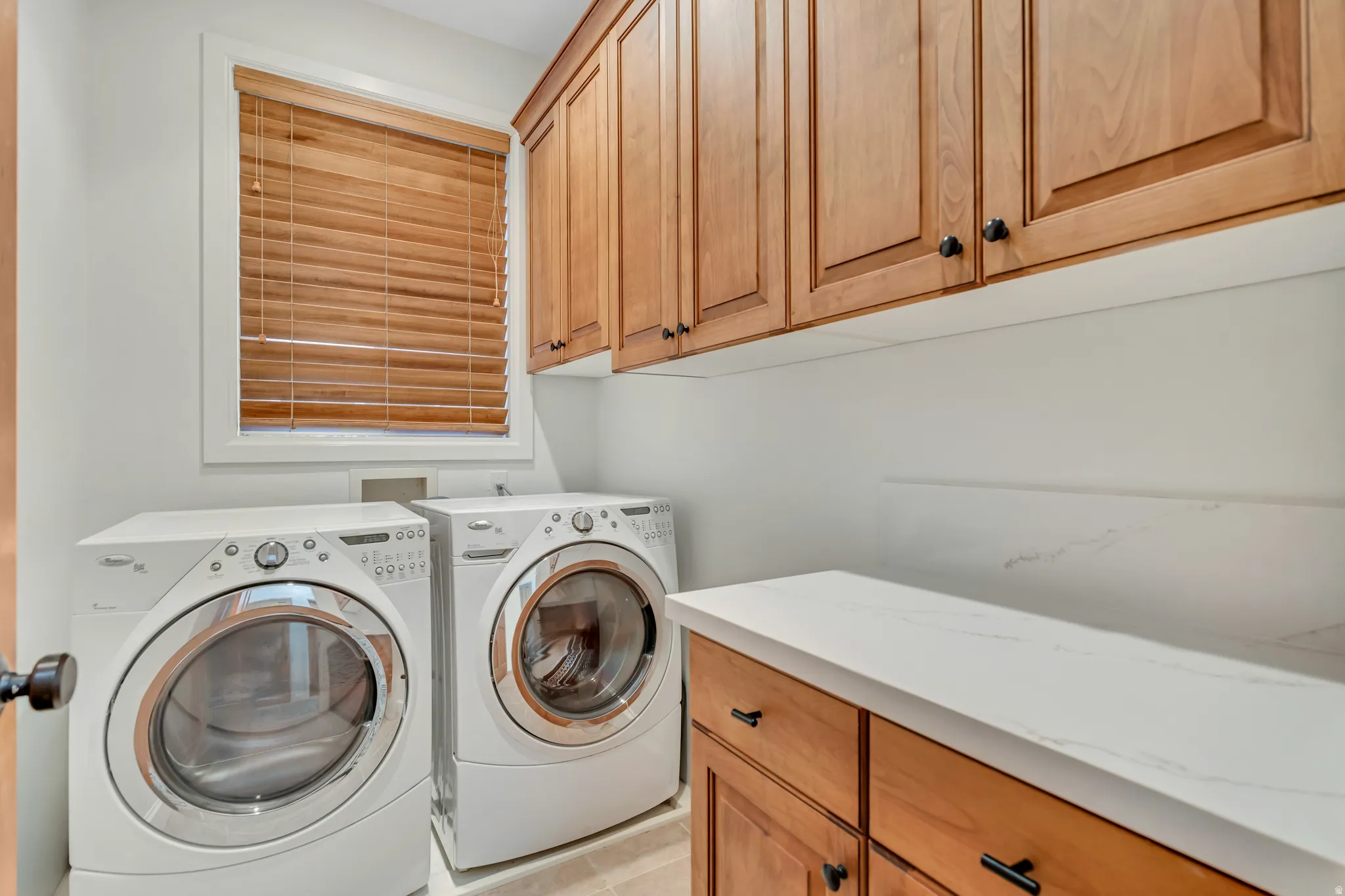 Dedicated laundry room featuring light wood cabinetry, white countertops, a window with wood blinds, and light-colored flooring