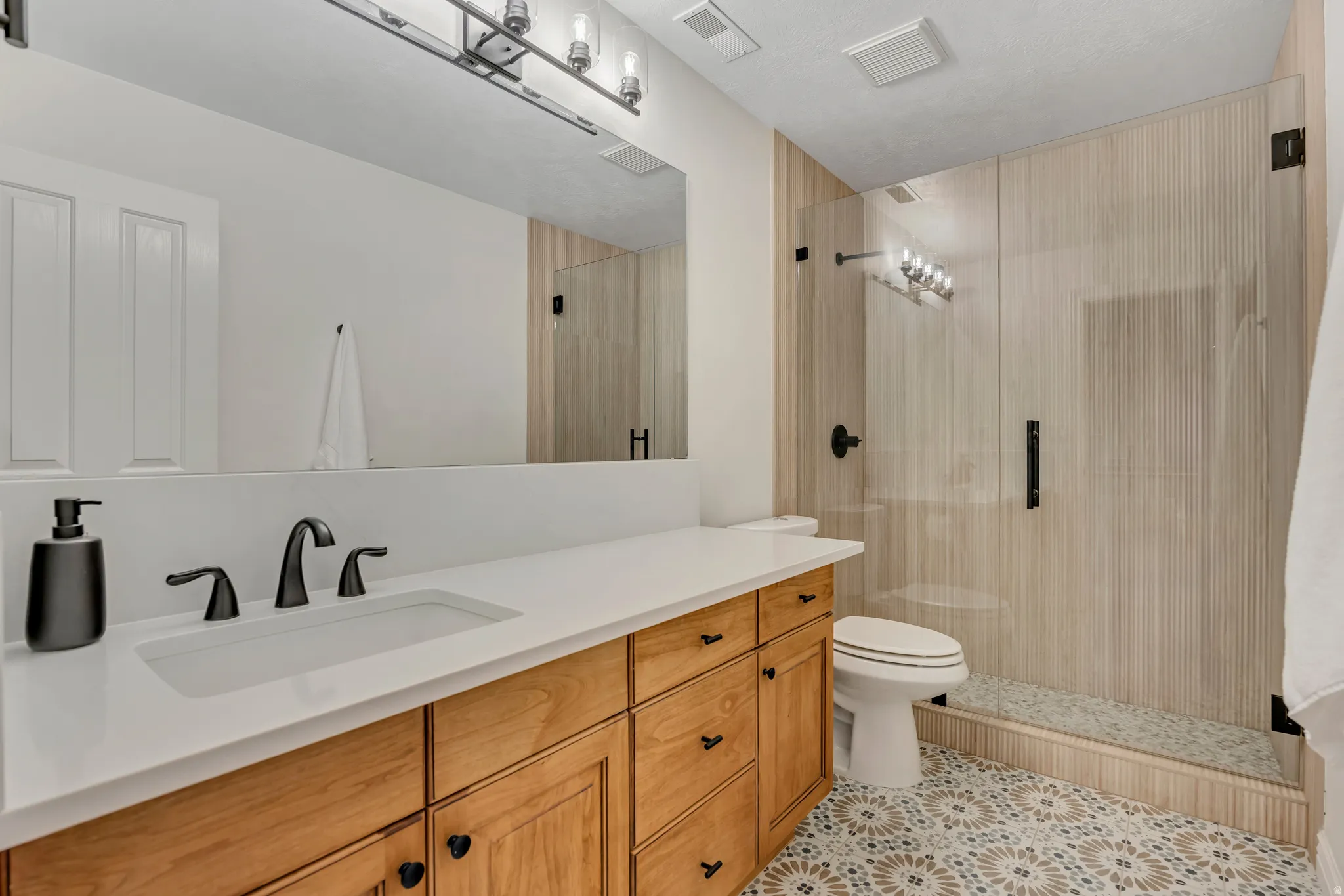 Bathroom featuring a wood-finish vanity with a white countertop and matte black fixtures, an undermount sink, a glass-enclosed shower with textured wall tile, and patterned floor tile