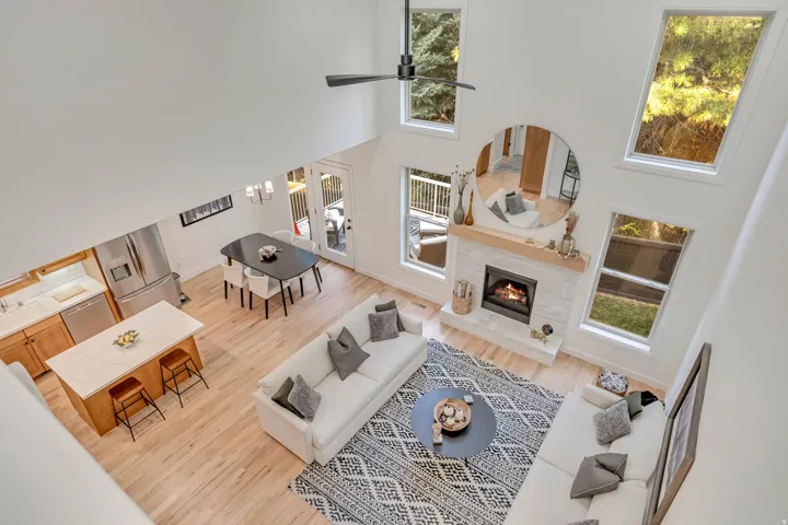 Open-concept living space featuring vaulted ceilings, abundant natural light from multiple windows, a modern fireplace with a light-toned tile surround and wood mantle, light wood-finish flooring, and a contemporary kitchen with a central island and stain