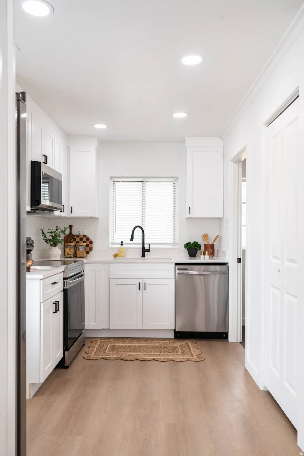 Galley kitchen featuring white cabinetry, light-toned wood-finish flooring, recessed lighting, stainless steel appliances, and a window with blinds