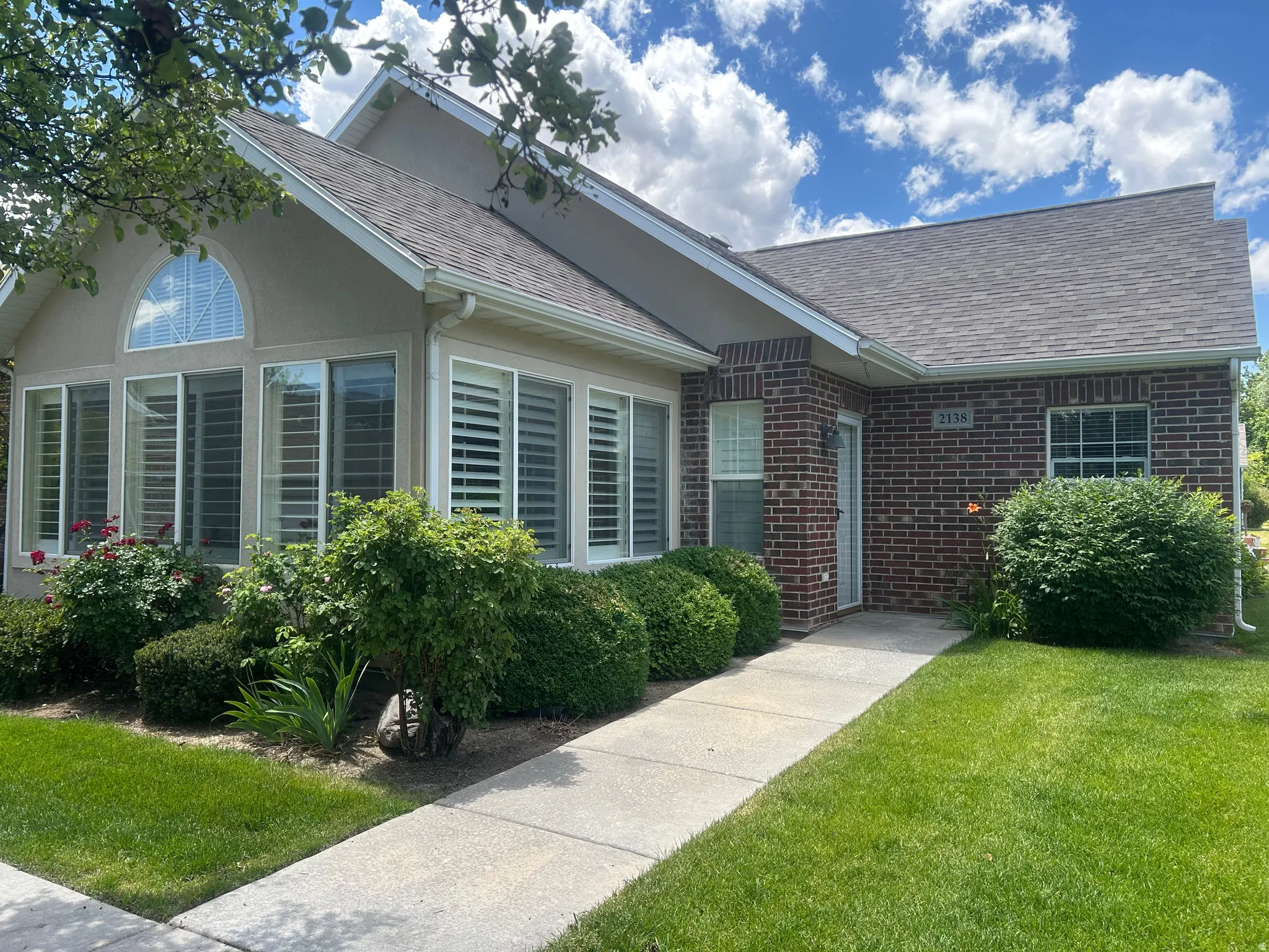 Exterior facade featuring brick and stucco finishes, multiple large windows with plantation shutters, an arched transom window, and a concrete pathway