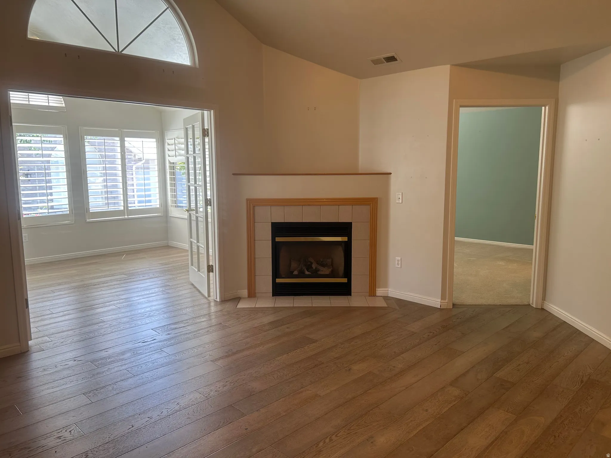 Living area featuring wood-finish flooring, a fireplace with a tiled surround, a half-moon transom window, and French doors leading to a sunroom with plantation shutters