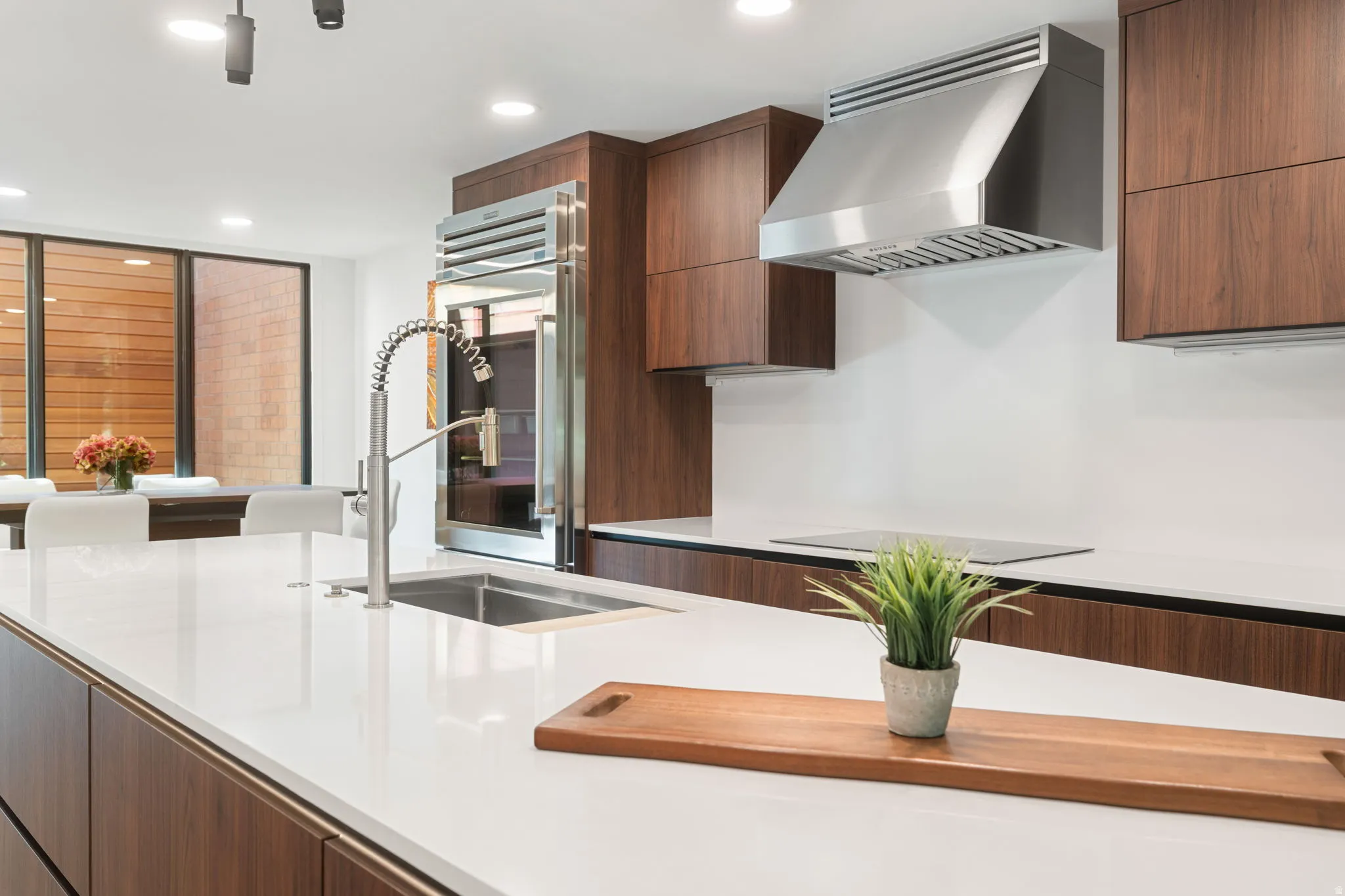 Kitchen featuring a large center island with integrated sink, stainless steel pull-down faucet, white solid surface countertops, wood-finish cabinetry, and stainless steel appliances including a built-in refrigerator and range hood
