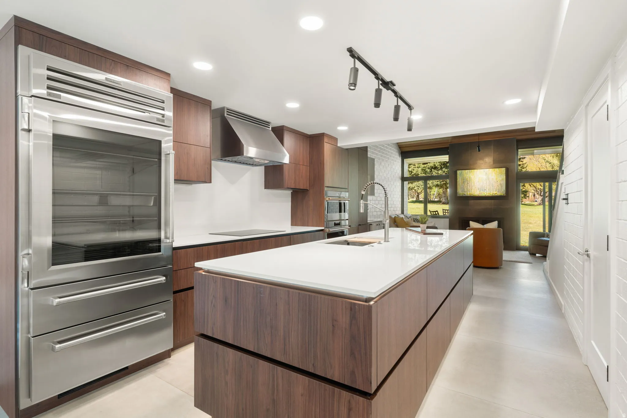 Contemporary kitchen featuring a large waterfall island with integrated sink, wood-finish cabinetry, stainless steel appliances, recessed lighting, and track lighting