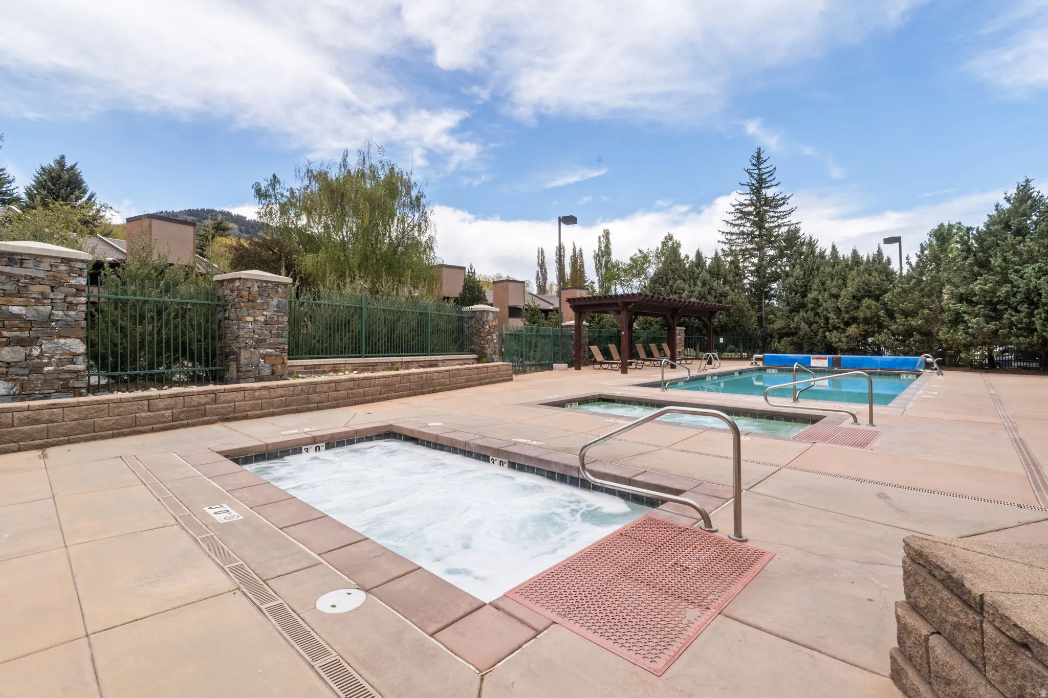 Outdoor spa and swimming pool area featuring a stone and block retaining wall, metal fencing, and a wooden pergola