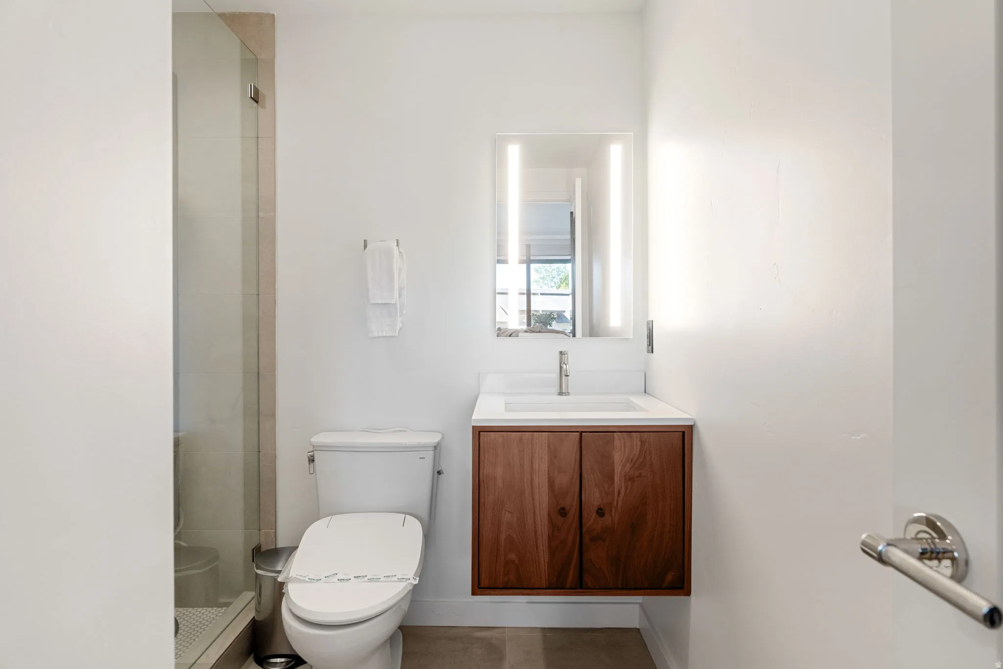 Modern bathroom featuring a floating wood-finish vanity with an integrated sink, a rectangular illuminated mirror, and a glass-enclosed shower with tile surround