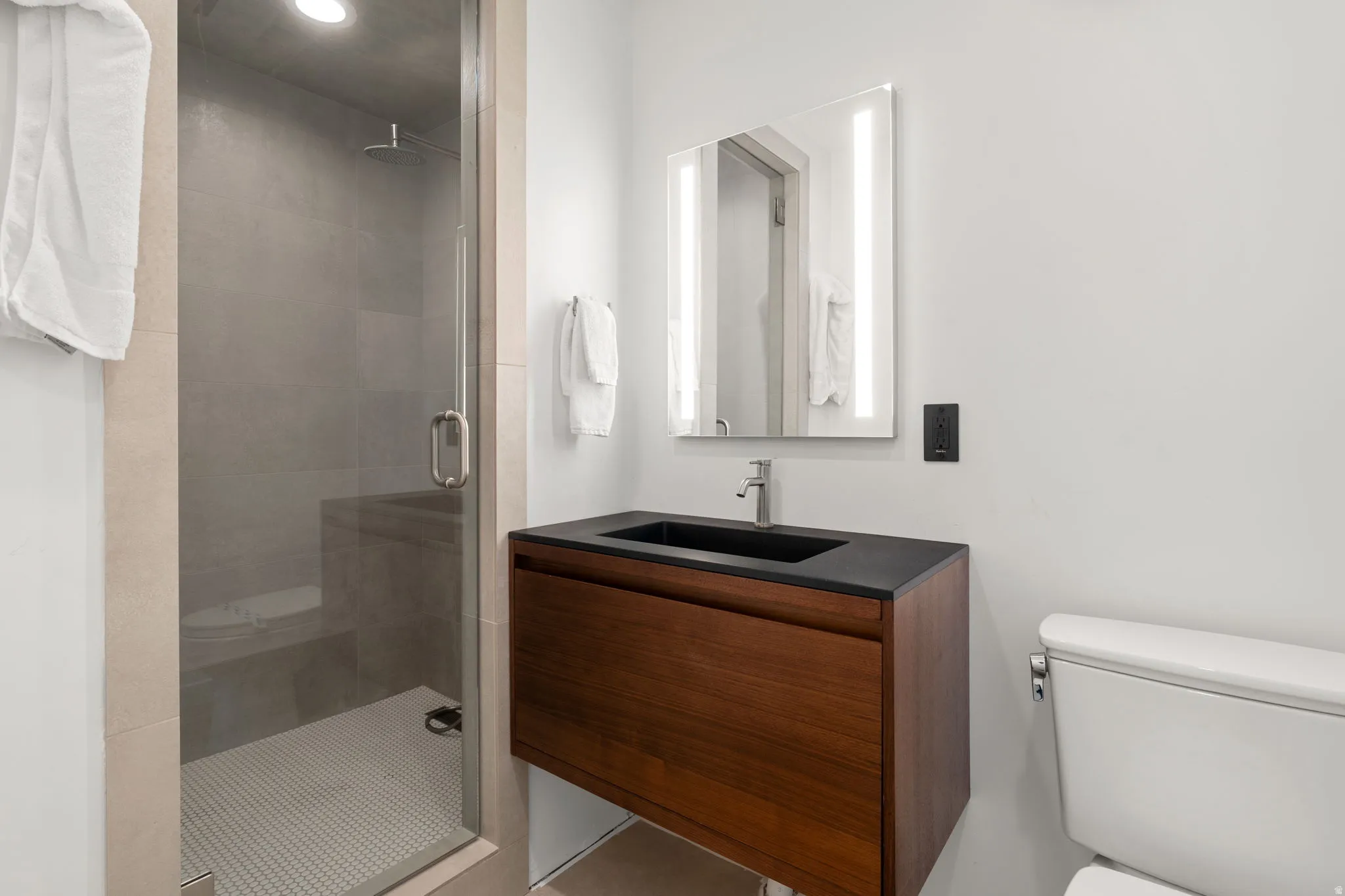 Bathroom featuring a glass-enclosed shower with a rain showerhead, a floating wood-finish vanity with an integrated black sink and stainless steel faucet, and an illuminated mirror
