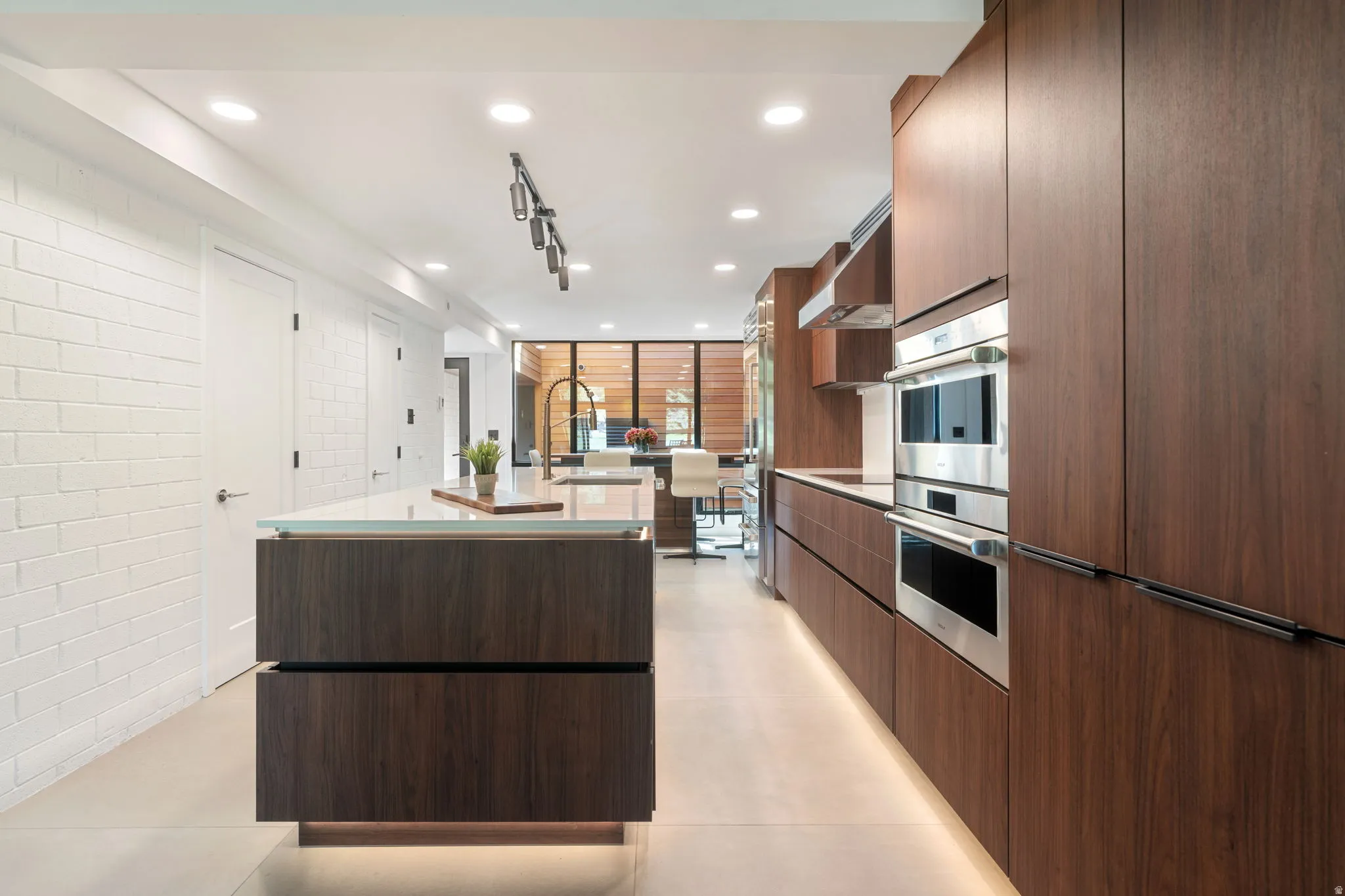Sleek kitchen featuring wood-finish cabinetry, integrated stainless steel appliances, a spacious island with a light-toned countertop, recessed lighting, and a white brick-textured wall
