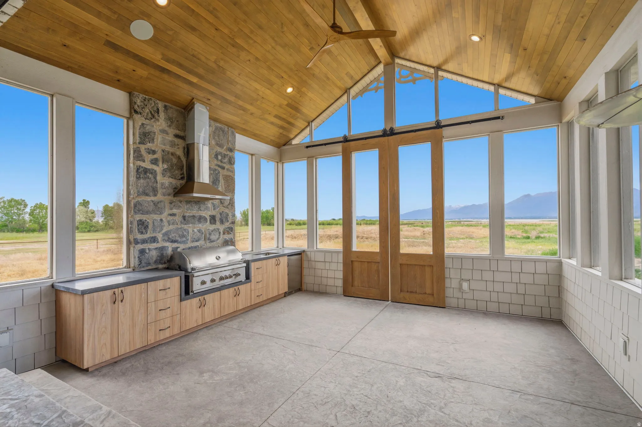 Expansive outdoor kitchen off the main kitchen featuring a built-in grill, stone accent wall, wood-finish cabinetry, vaulted wood-plank ceiling, and large windows with sliding barn doors
