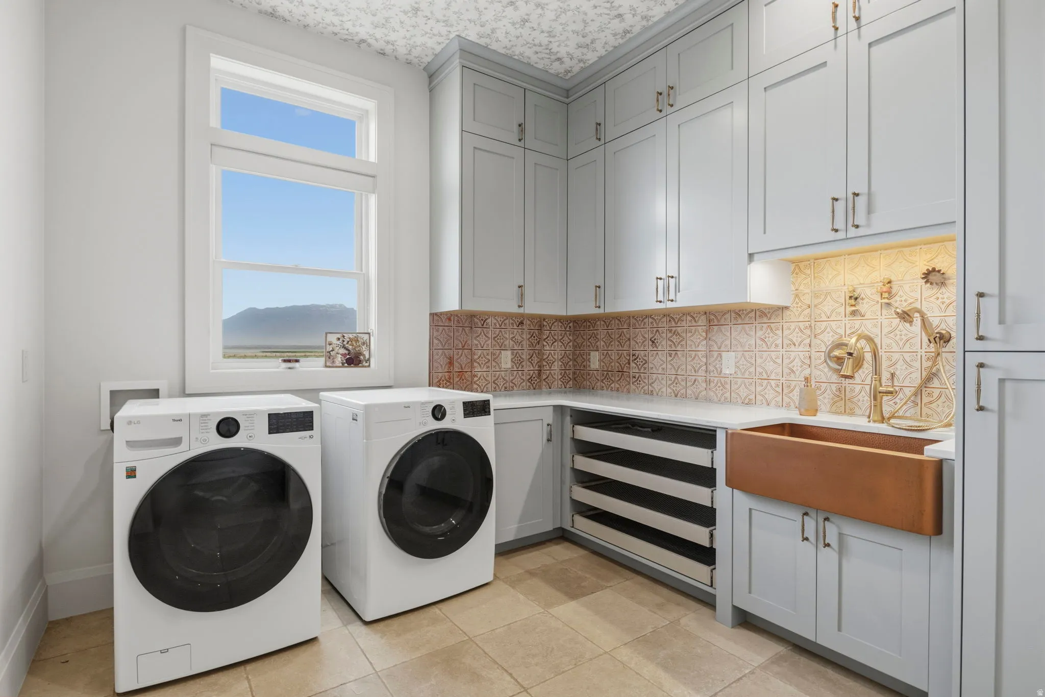 Dedicated laundry room featuring light gray cabinetry, a copper farmhouse sink with brass fixtures, textured tile backsplash, ample counter space, and integrated pull-out drying racks