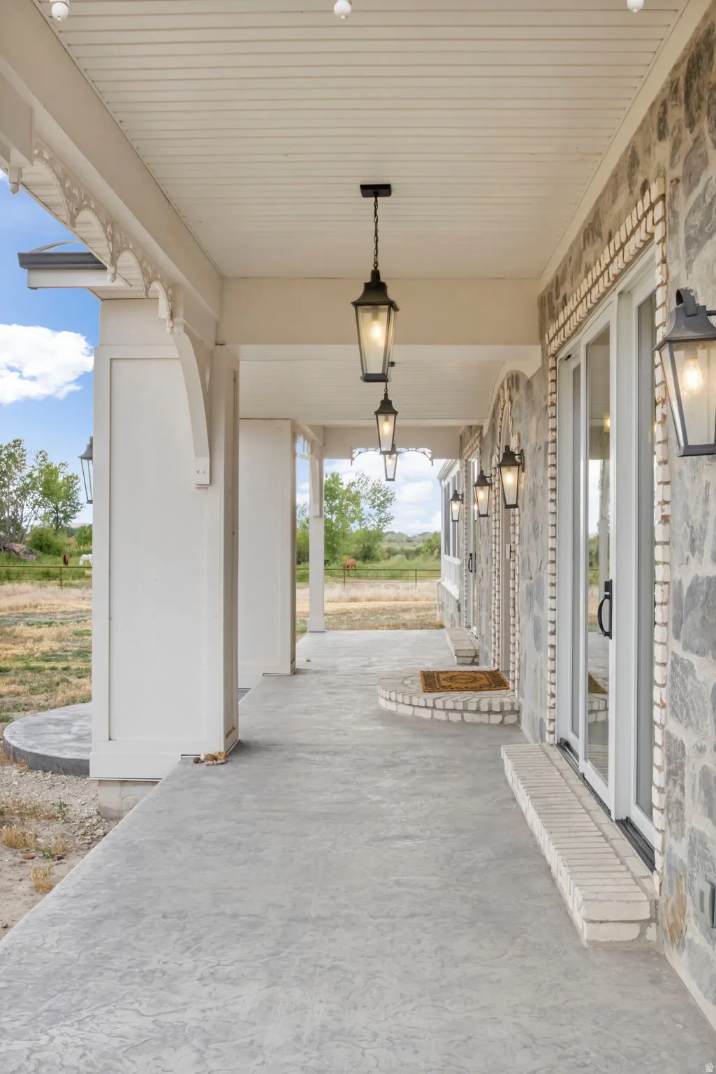 Covered patio featuring stamped concrete flooring, a stone facade, white columns with decorative corbels, and black pendant lighting fixtures
