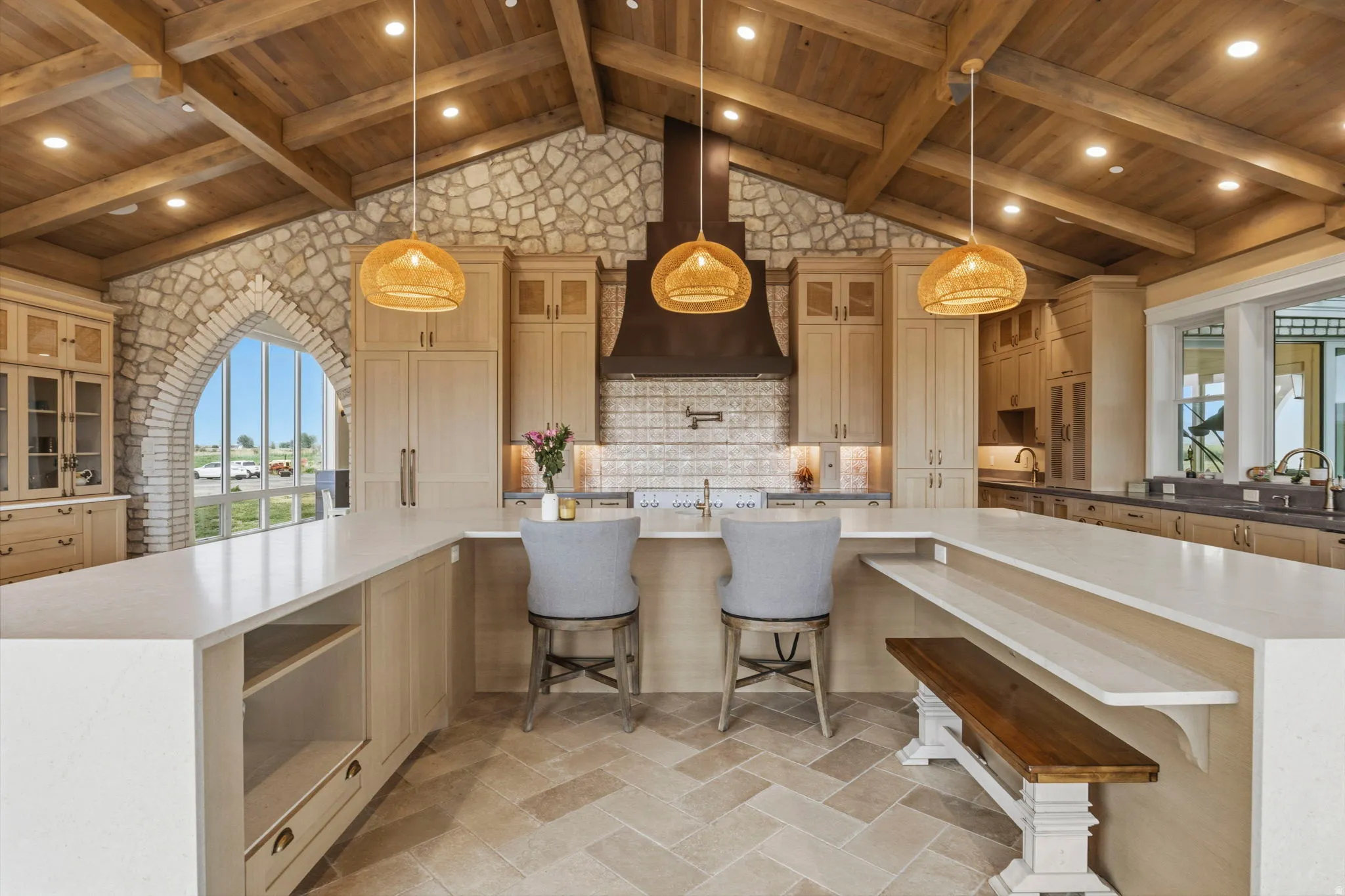 Expansive kitchen featuring a vaulted wood-beamed ceiling, stone accent walls, a large island with integrated seating, light-toned cabinetry, and herringbone-patterned tile flooring