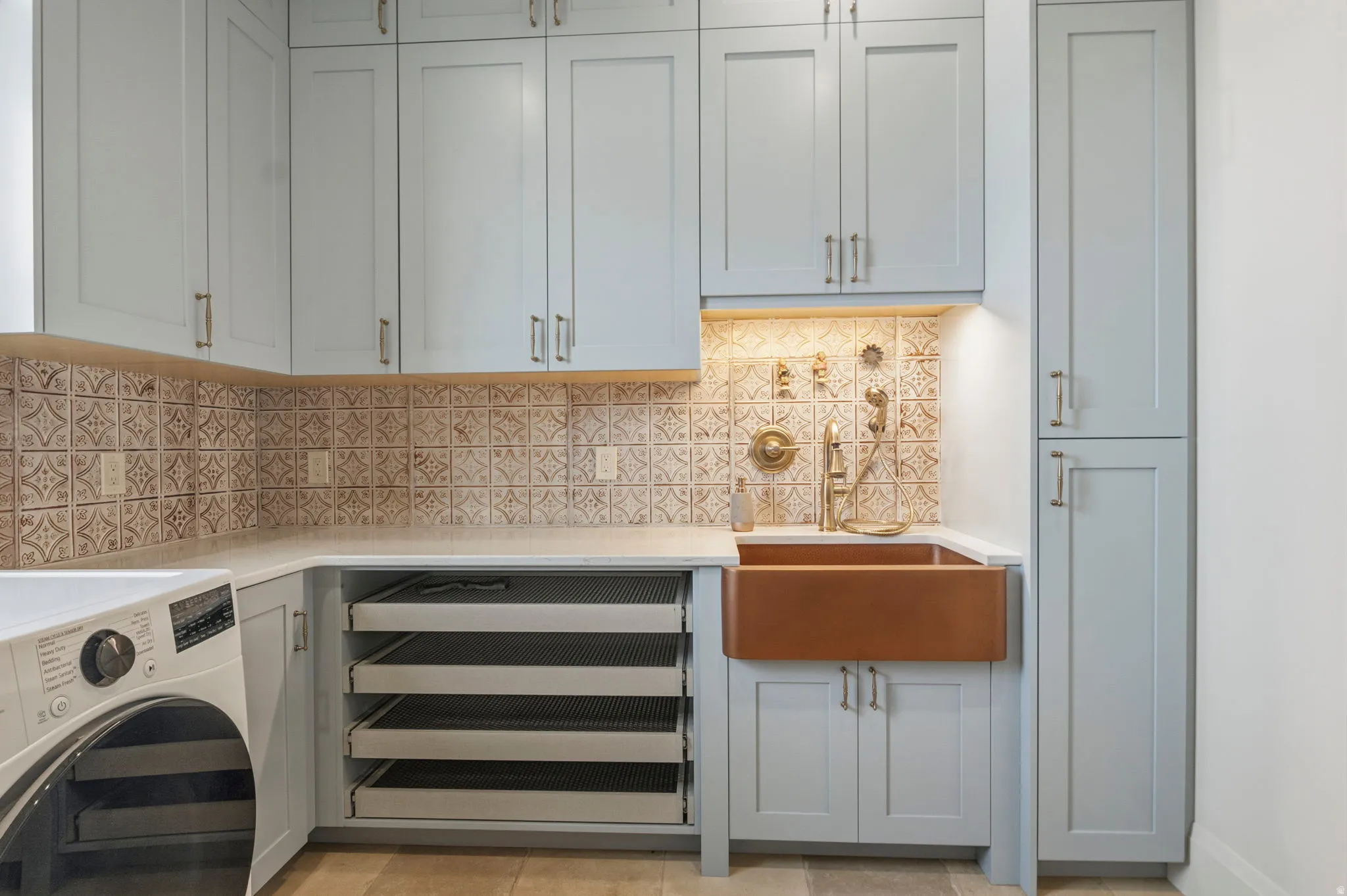 Utility room featuring custom light blue cabinetry, a large farmhouse-style copper sink which doubles as a dog wash with brass fixtures, under-cabinet lighting, and a patterned tile backsplash