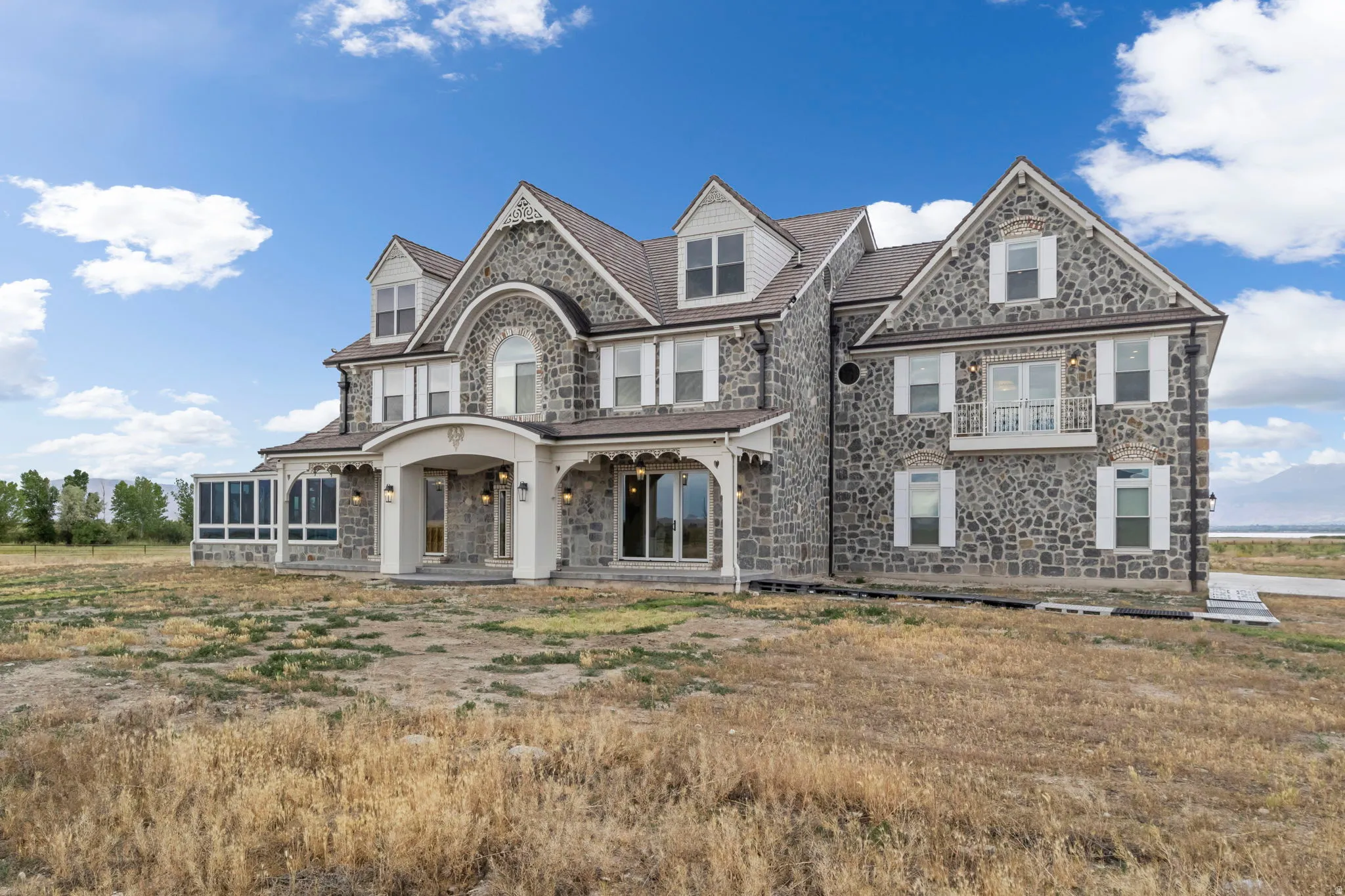 Stone-clad exterior featuring a prominent arched entrance with columns, a covered porch, multiple dormer windows, a sunroom extension, and a second-story balcony