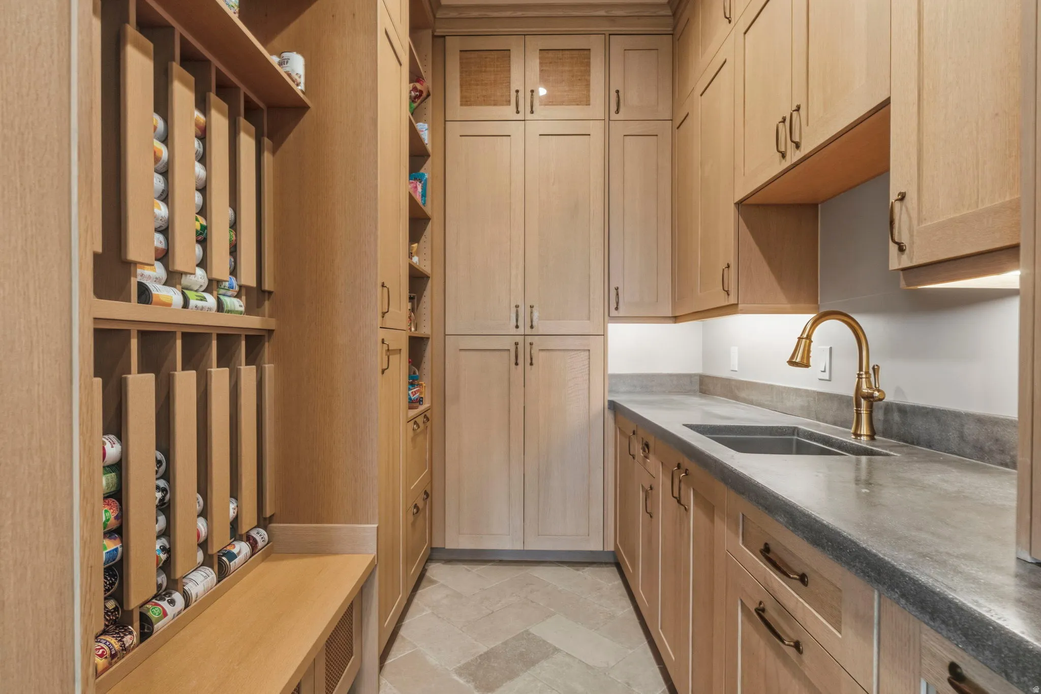 Custom pantry featuring light wood cabinetry, integrated roll-out storage, durable countertops, and an undermount sink with a bronze-finish faucet