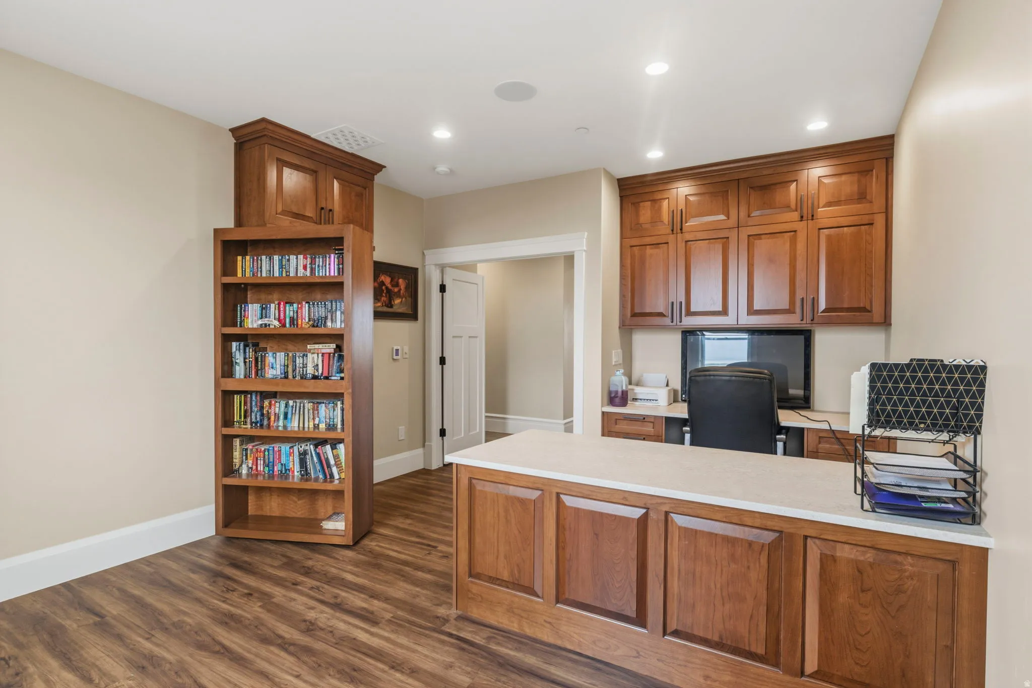 Dedicated workspace featuring wood-finish flooring, extensive built-in cabinetry, recessed lighting, and a substantial built-in desk with a light-toned countertop. Secret bookshelf door to workshop