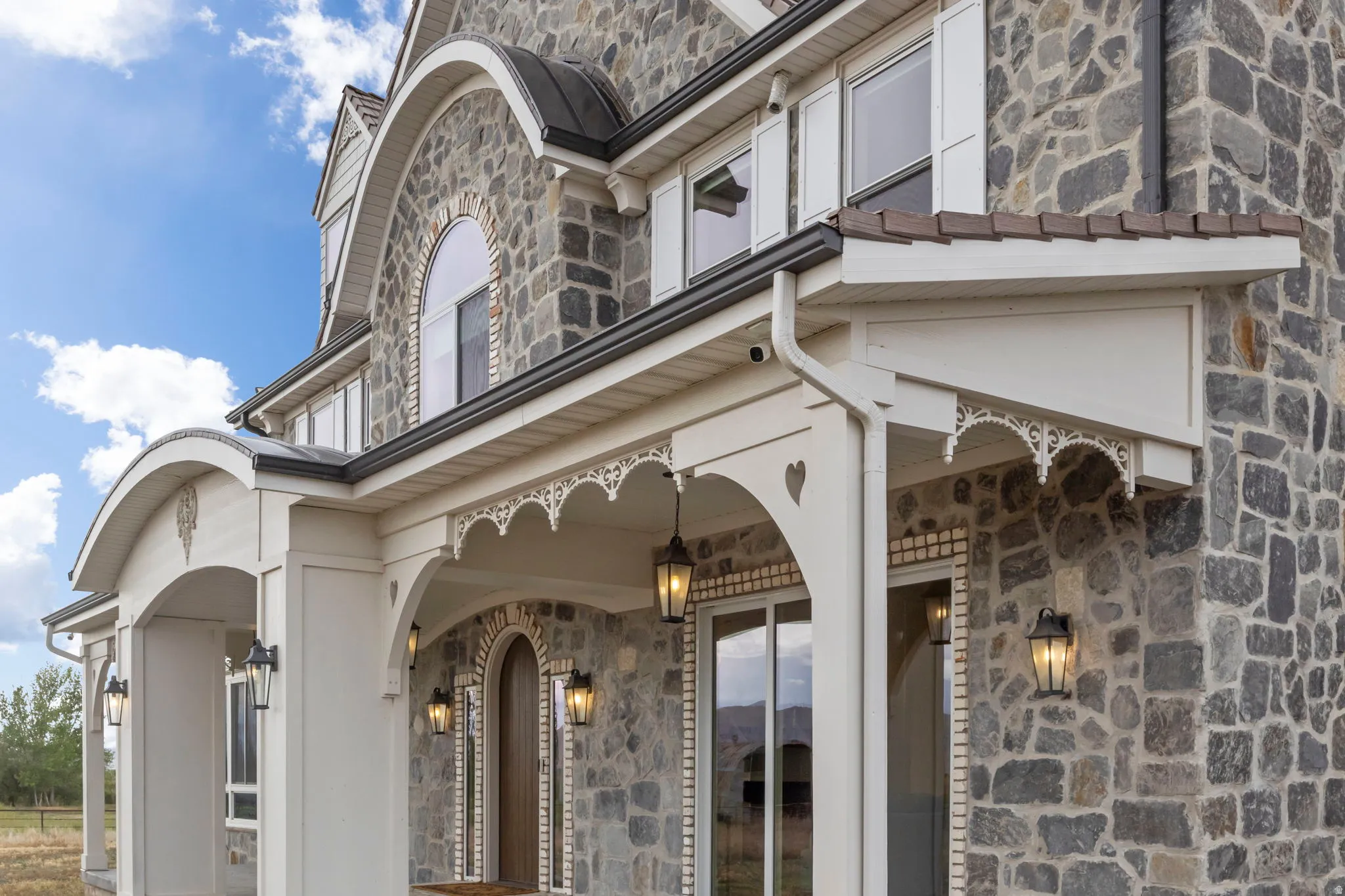 Stone facade featuring a covered porch with intricate gingerbread trim, arched entryway, wall-mounted lanterns, and white window shutters