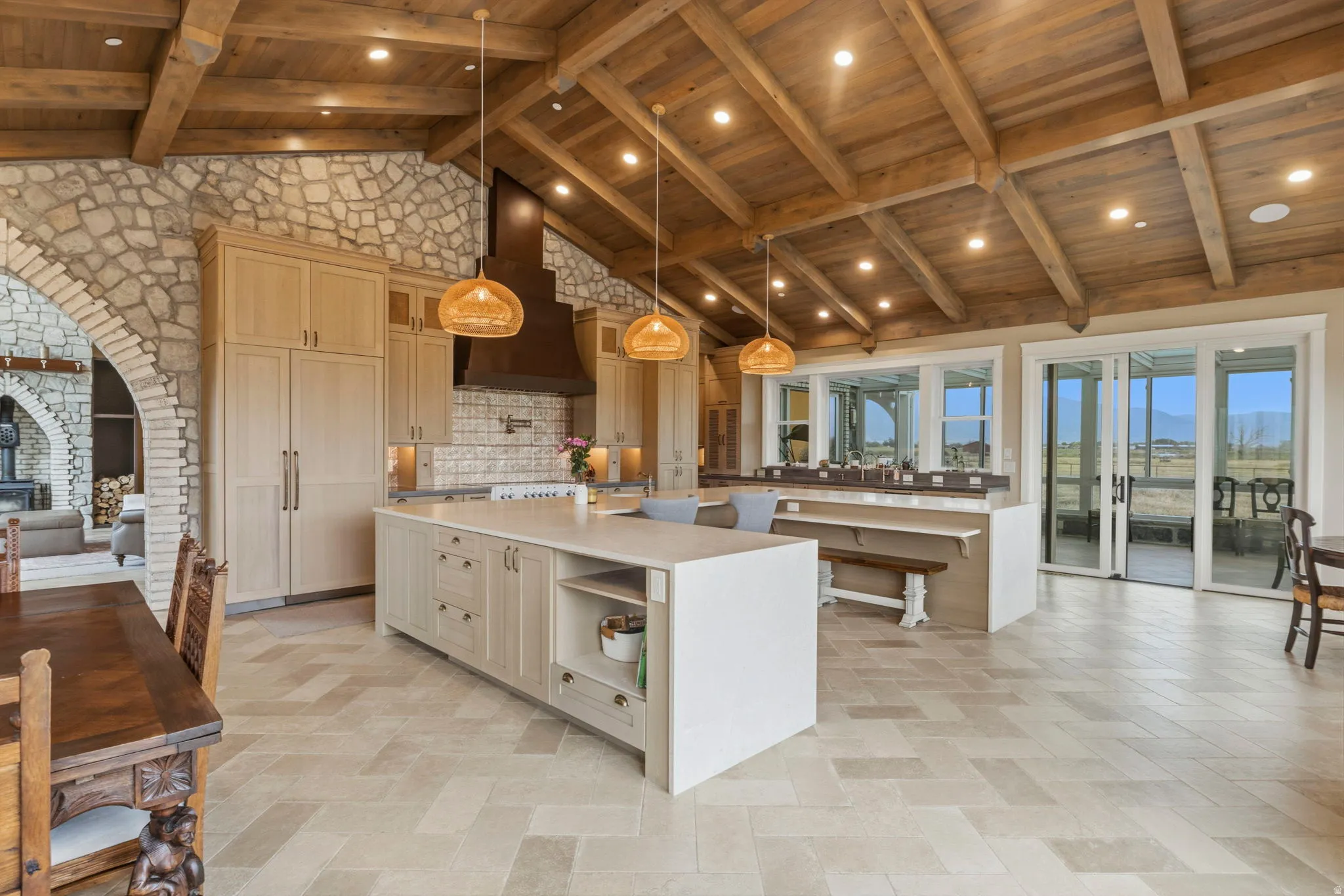 Gourmet kitchen featuring a vaulted wood-beam ceiling, stone accent walls, custom cabinetry with integrated refrigerator, a large center island with seating, and expansive sliding glass doors
