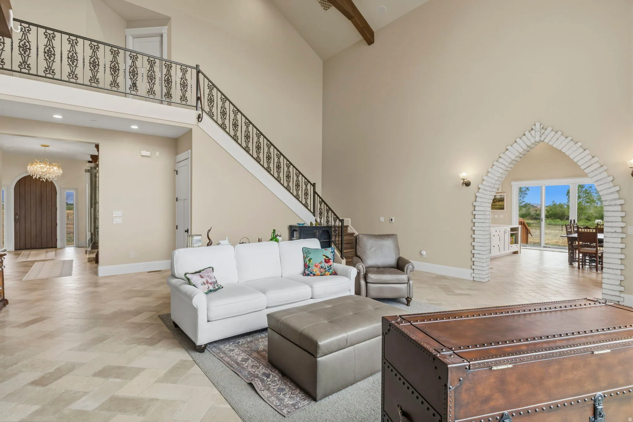 Spacious living area featuring high ceilings, exposed wood beam, herringbone-patterned flooring, a decorative iron staircase, and an arched brick entryway