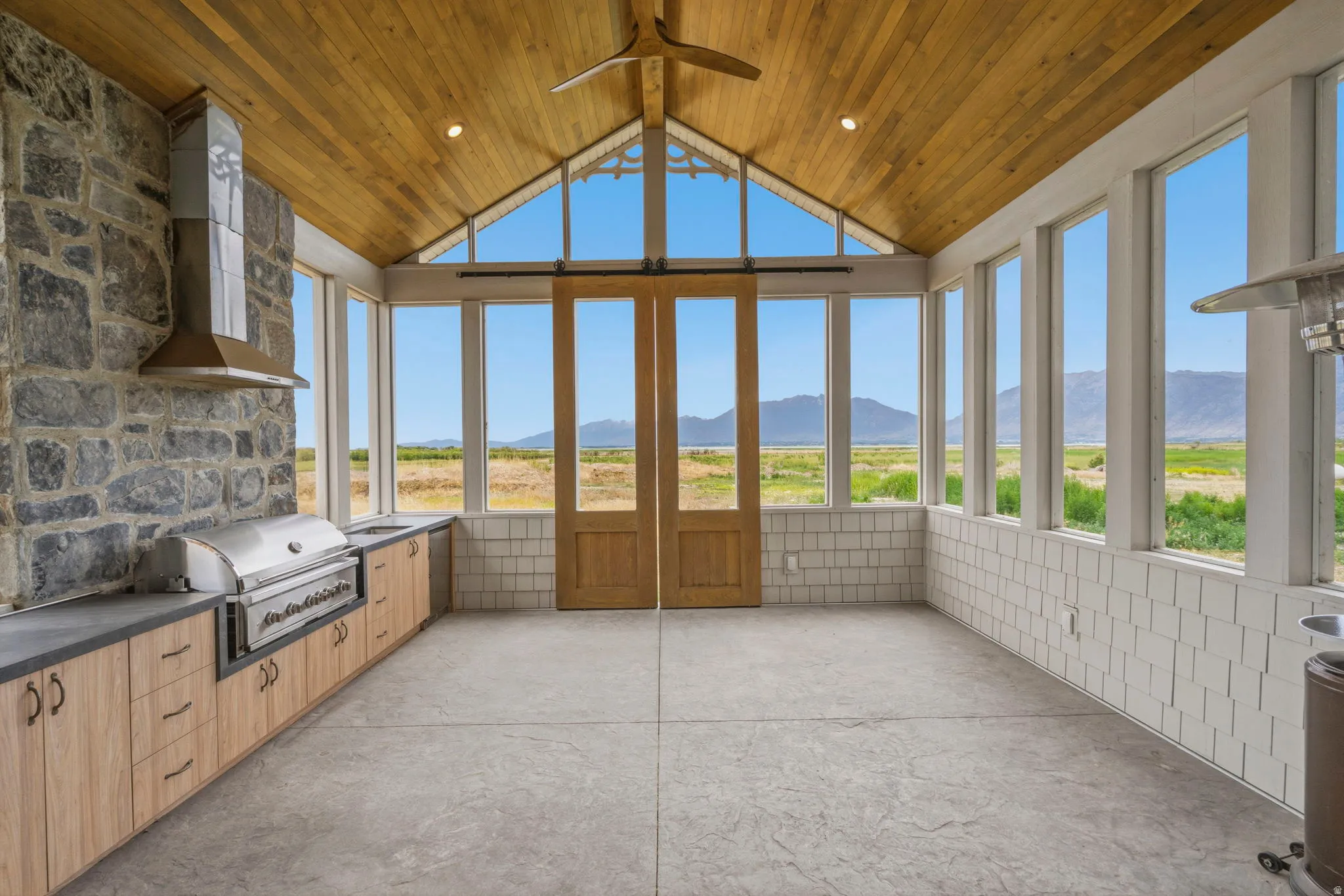 Expansive outdoor kitchen with enclosed patio featuring a vaulted wood-paneled ceiling, full-height stone accent wall, and integrated outdoor kitchen with built-in grill and range hood. Barn doors open to enjoy seasons with that indoor outdoor feel.