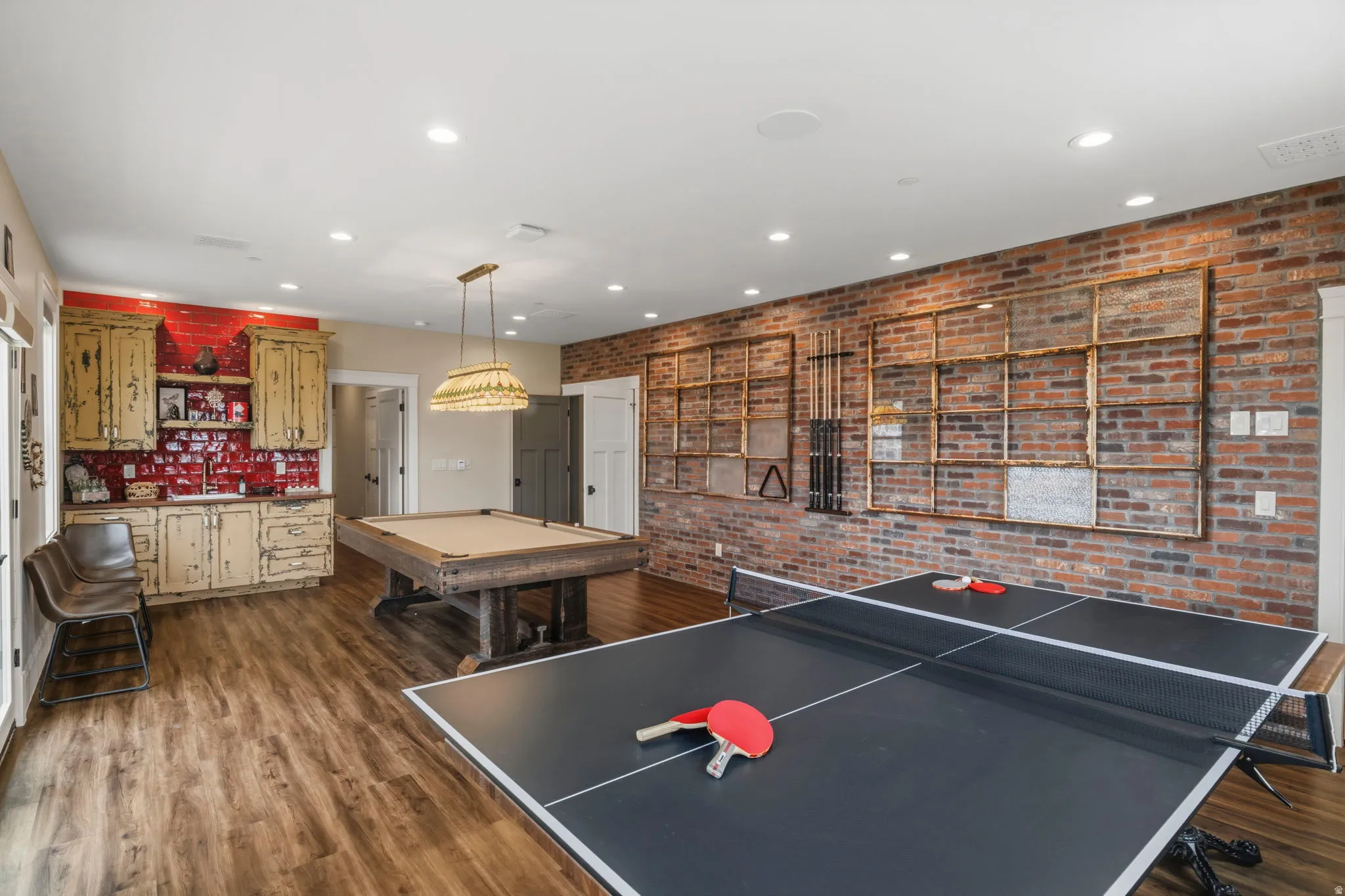 Spacious entertainment area featuring wood-finish flooring, recessed lighting, and a brick accent wall with decorative shelving