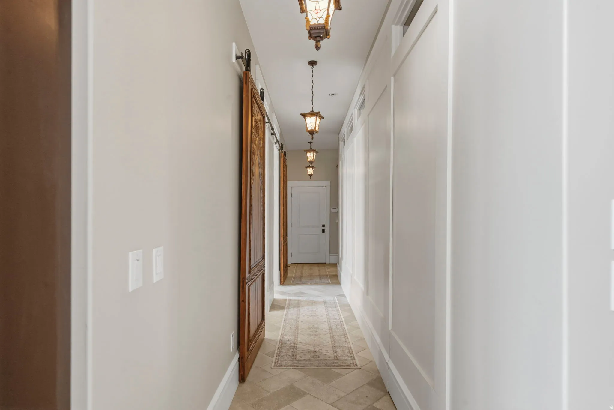 Hallway featuring natural wood barn doors, decorative pendant lighting, tile flooring with an area rug, and white wall paneling, one of which is a secret door.