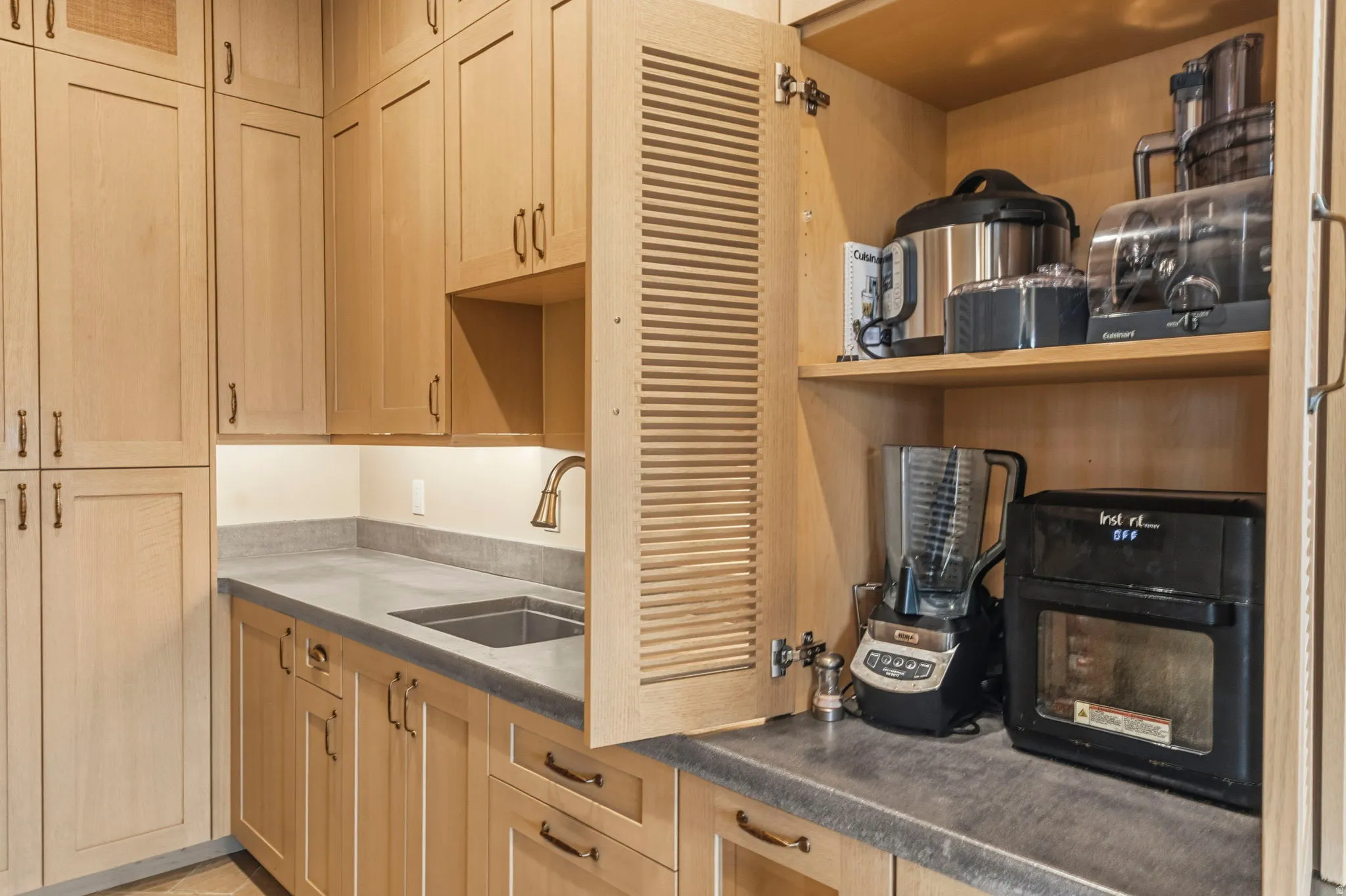 Built-in appliance garage cabinetry with natural wood-finish doors and bronze hardware, integrated sink with gooseneck faucet, grey countertop, and under-cabinet lighting