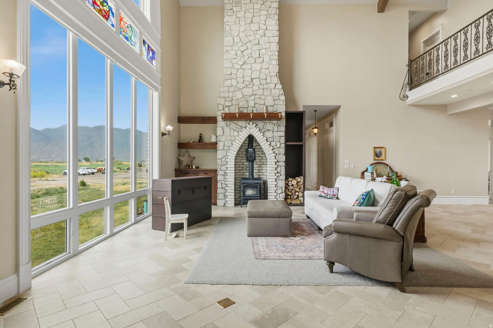Spacious living area featuring floor-to-ceiling windows, a stone fireplace with a wood-burning stove, and a second-story balcony with decorative ironwork