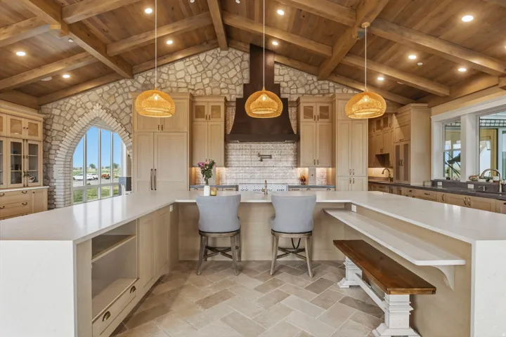 Expansive kitchen featuring a vaulted wood-beamed ceiling, stone accent walls, a large island with integrated seating, light-toned cabinetry, and herringbone-patterned tile flooring