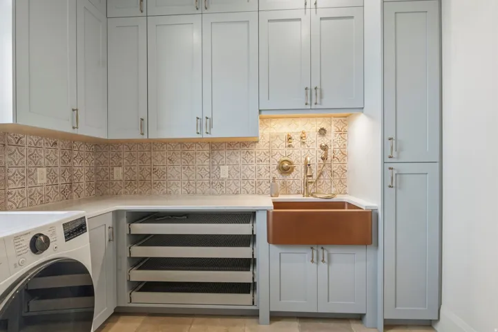 Utility room featuring custom light blue cabinetry, a large farmhouse-style copper sink which doubles as a dog wash with brass fixtures, under-cabinet lighting, and a patterned tile backsplash