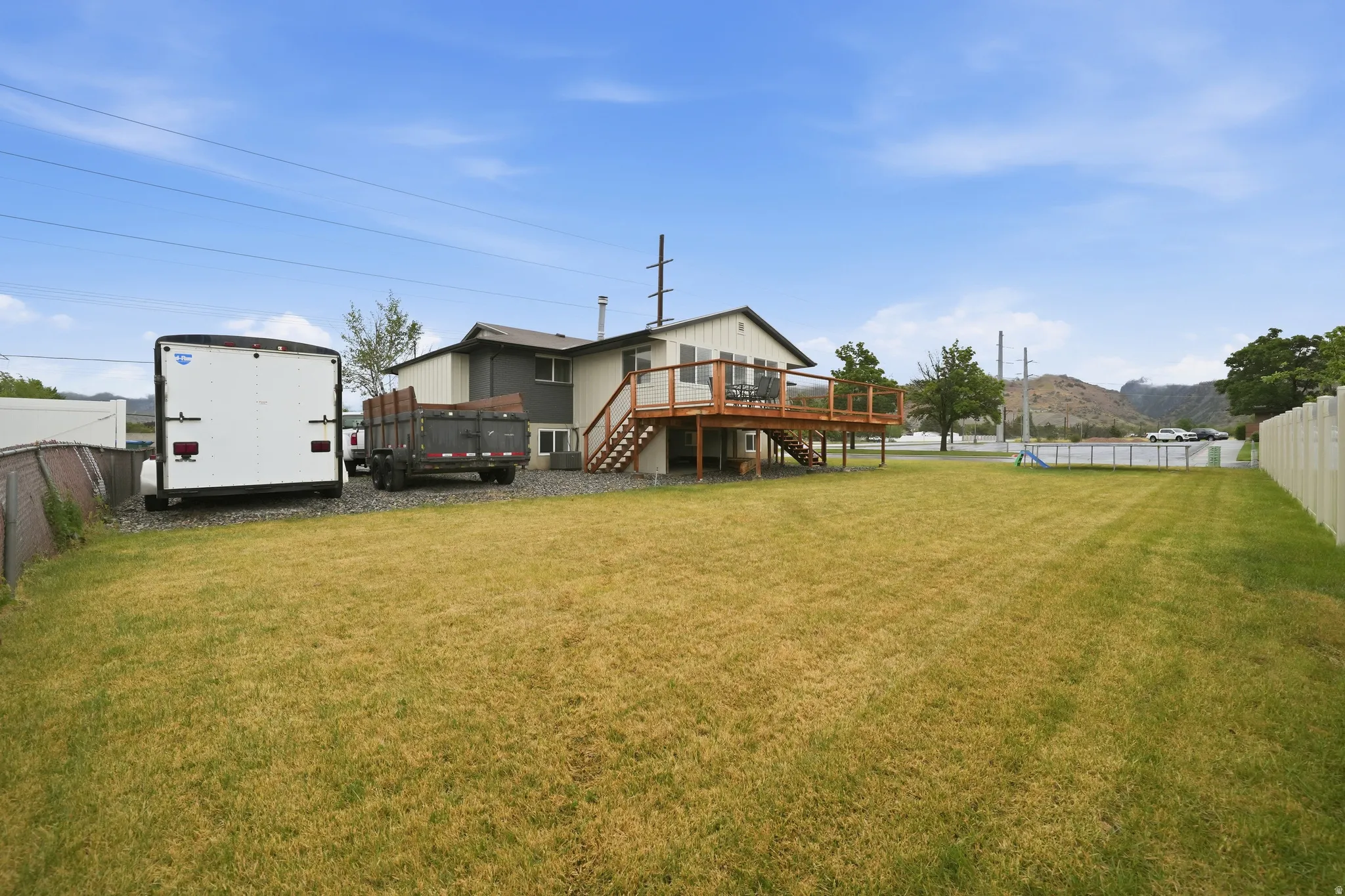 Expansive backyard featuring a large grassy area, a raised wood deck with staircase, and a gravel parking pad