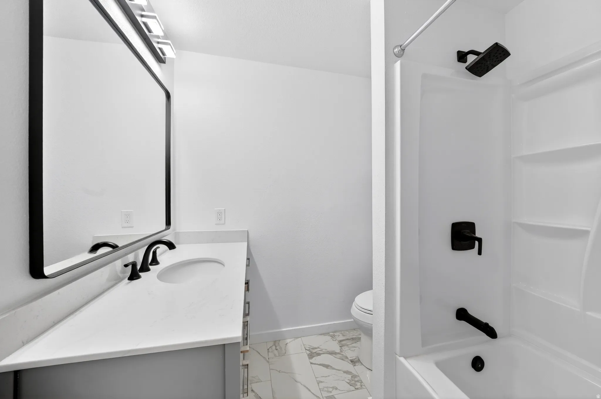 Bathroom featuring a white bathtub/shower combination with matte black fixtures, a single vanity with a white countertop and integrated sink, a rectangular framed mirror, and marble-finish floor tiling