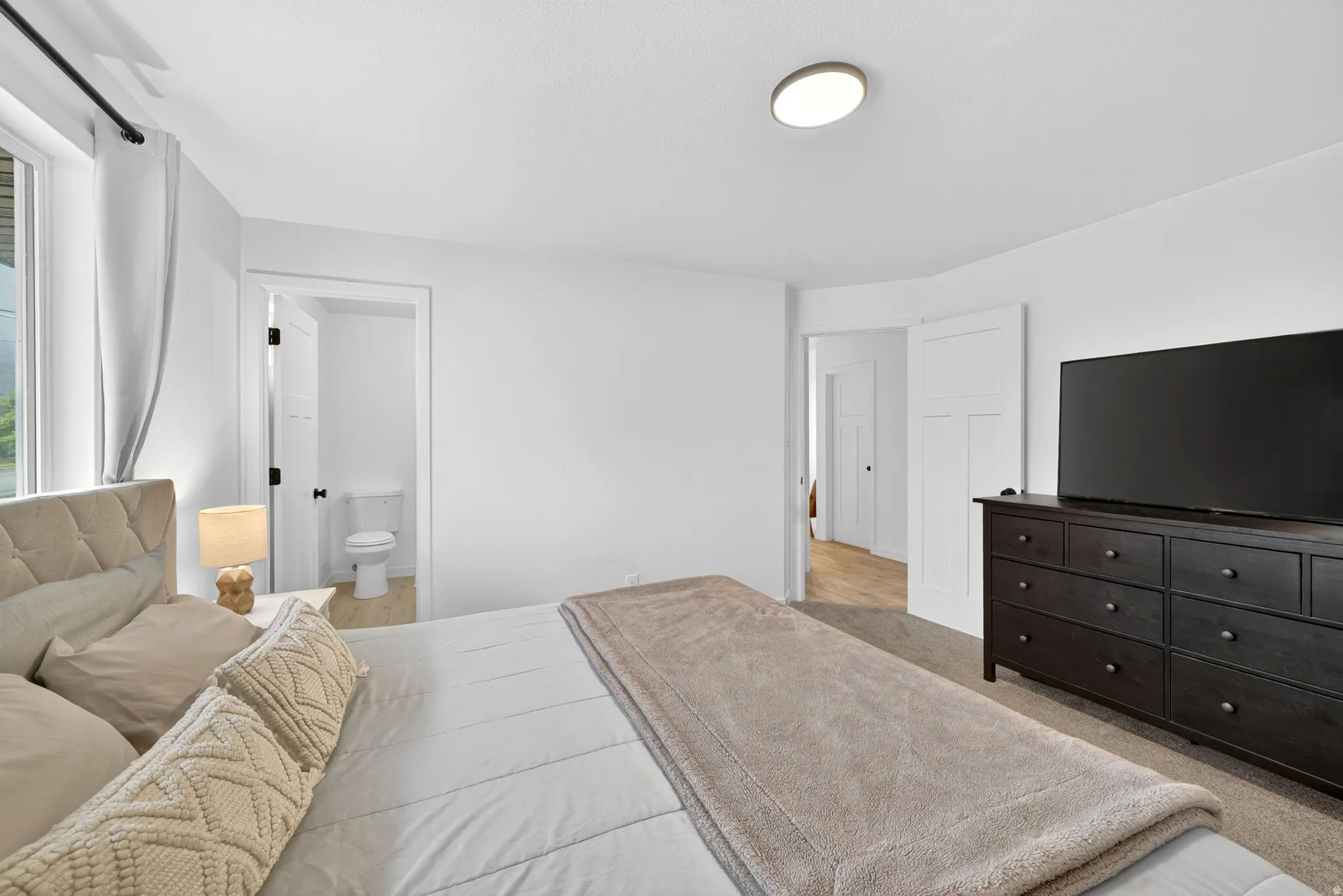 Carpeted bedroom featuring a built-in overhead light fixture, white walls, and a window with white drapes