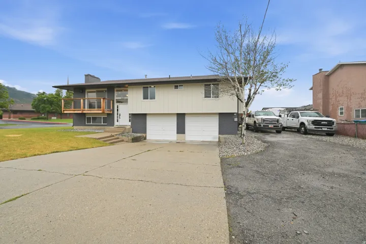 Split-level residence featuring a two-car garage, an upper-level balcony with wood railing, a paved driveway, and a gravel side parking area